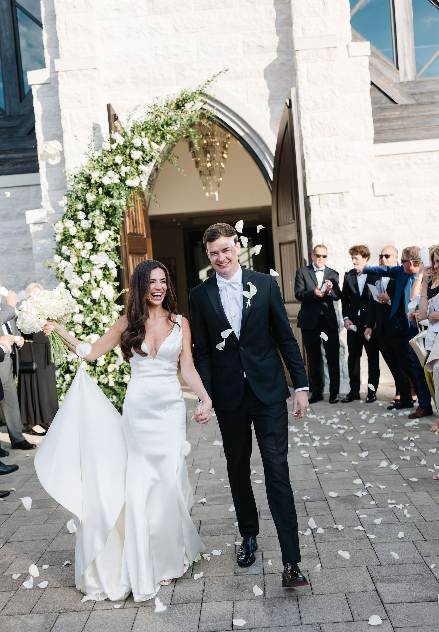 A happy couple exits a church through an arched doorway adorned with white floral arrangements as guests shower them in confetti.