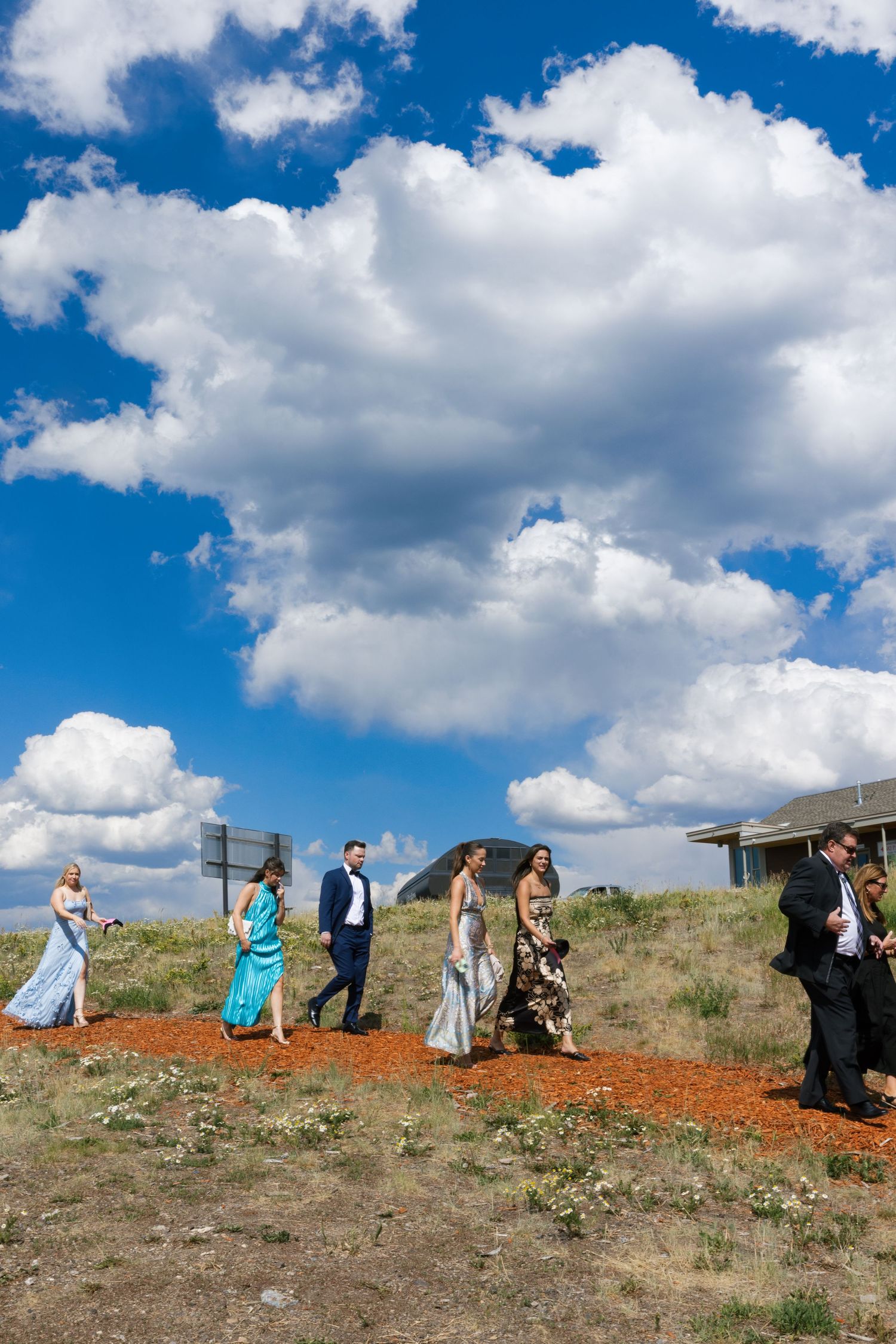 A group of people in formal attire walk along a hilltop ridge against a bright blue sky with white puffy clouds.