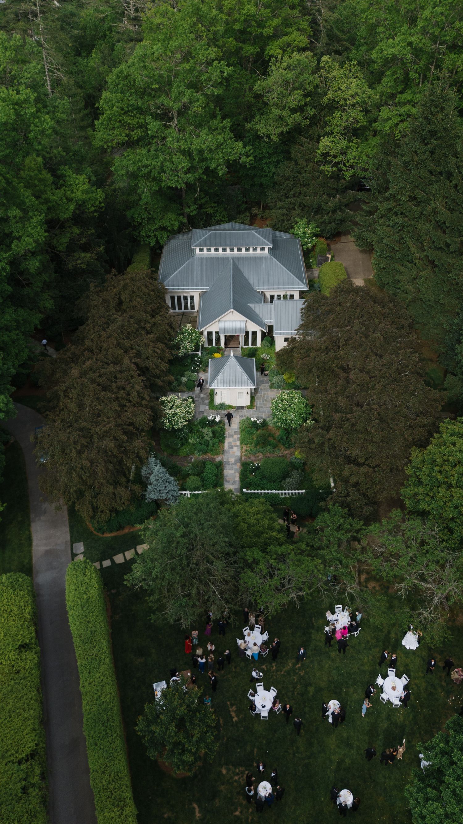 Aerial view of a white house with a garden path surrounded by lush trees and people gathered on the lawn for an event.