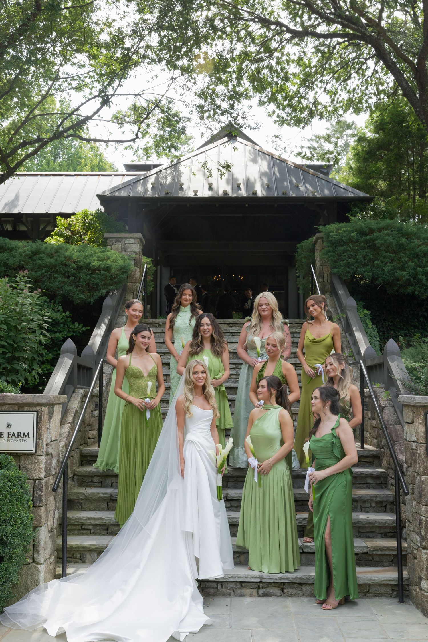 Bride in a white gown with a long veil stands before bridesmaids in green dresses on stone steps outside a rustic wooden pavilion.