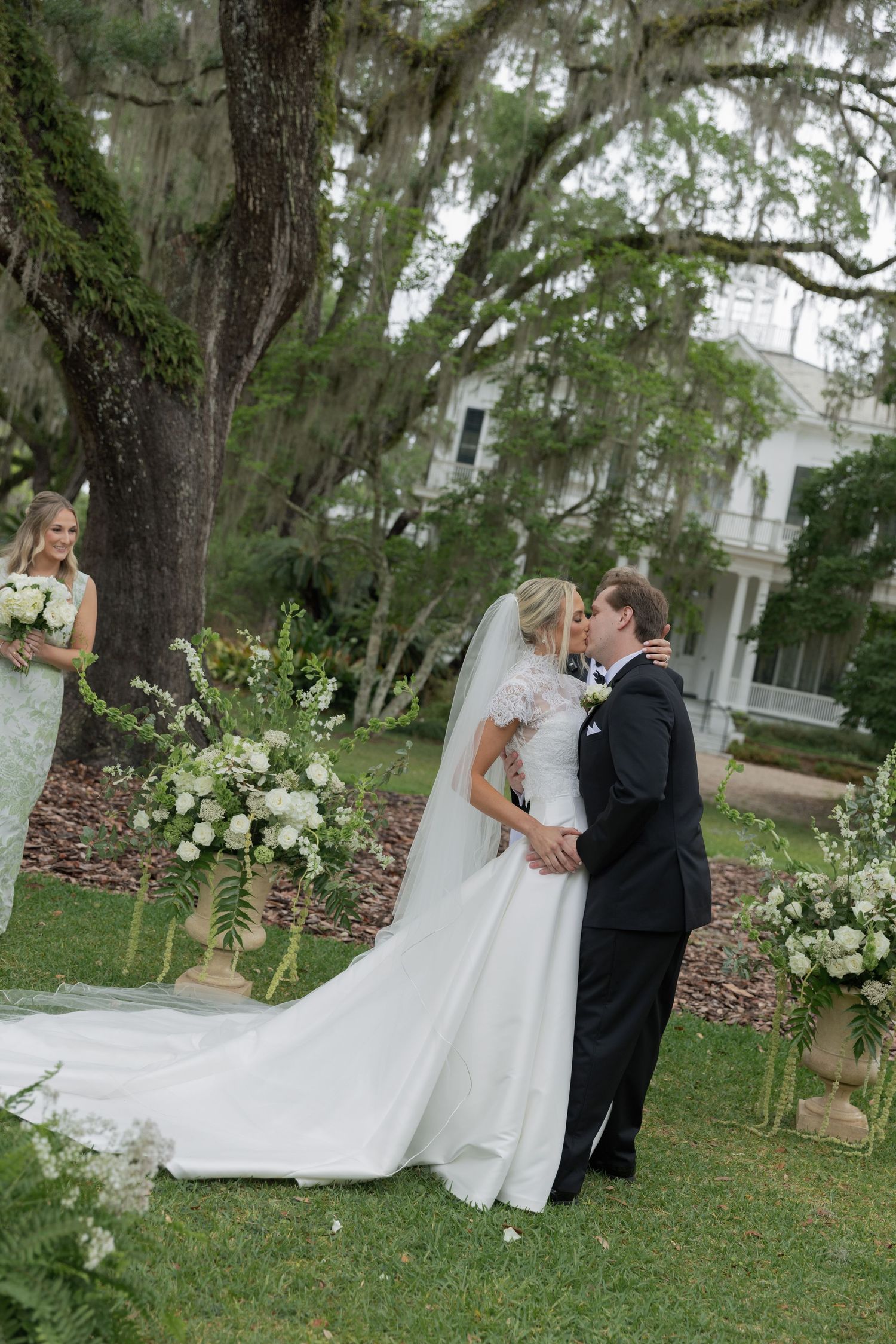 Bride and groom share first kiss outdoors under Spanish moss trees at elegant Southern plantation wedding ceremony.