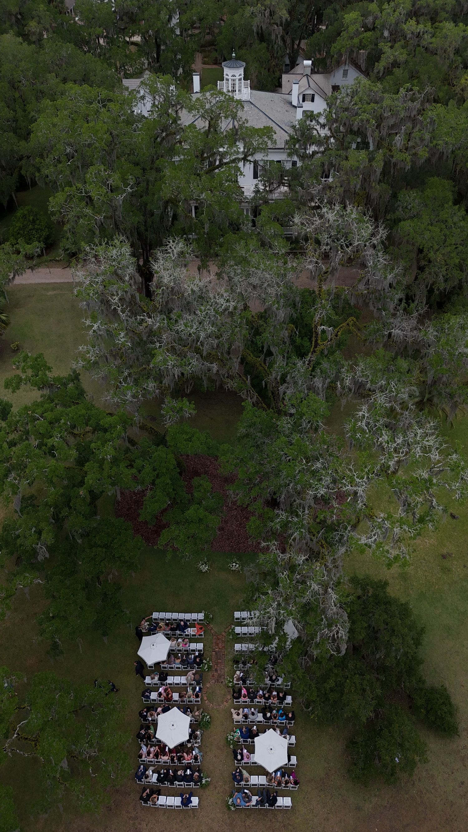 Aerial drone view of an outdoor wedding reception with white umbrellas on a lawn beside a historic Southern mansion surrounded by oak trees.