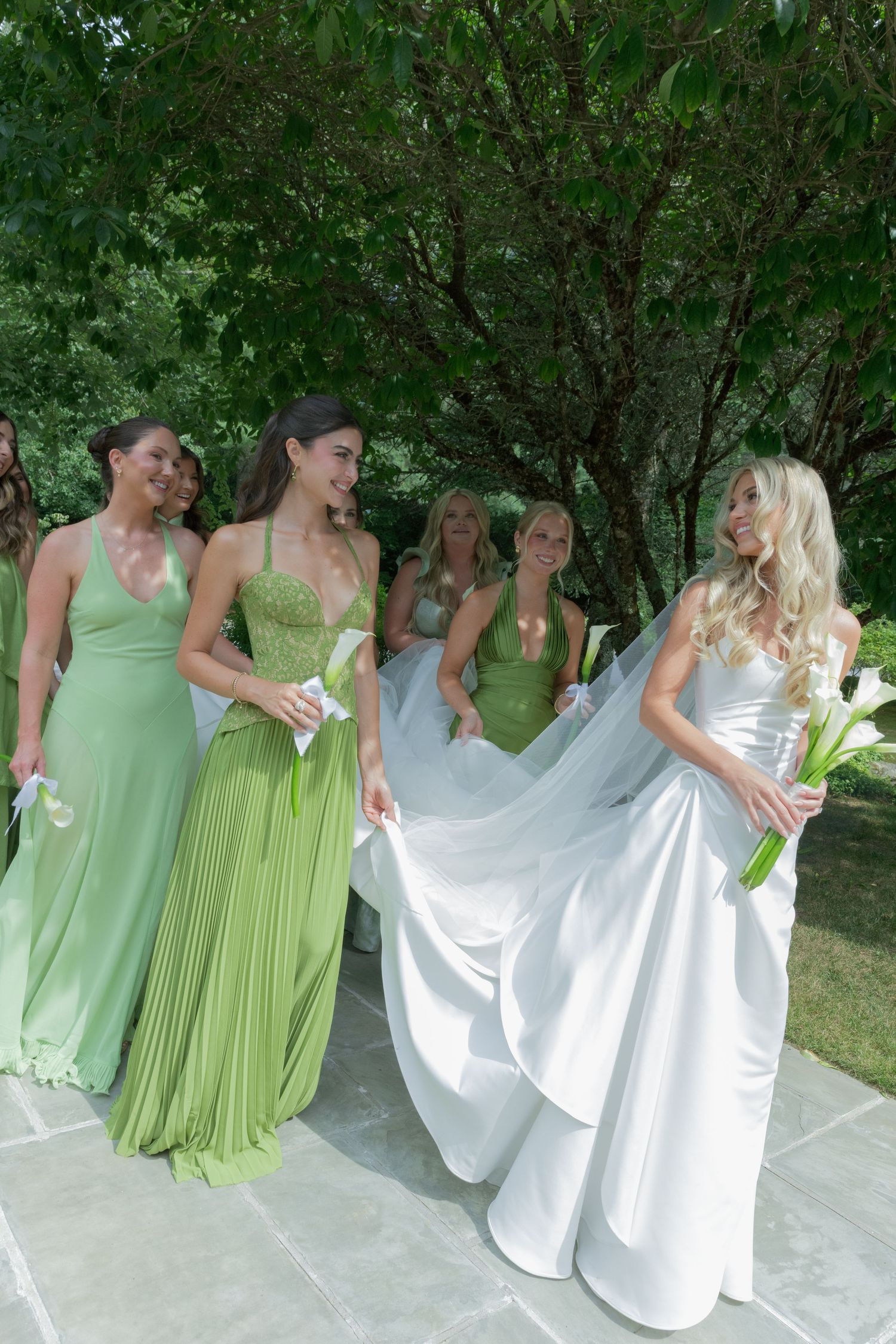 Bride in a flowing white satin gown with a long veil walks beside bridesmaids in green dresses holding white calla lilies outdoors.