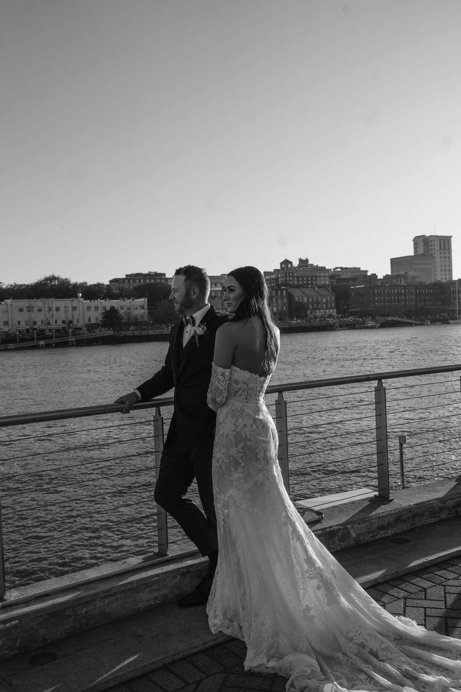 Black and white photo of bride and groom walking on waterfront dock with city skyline in background.