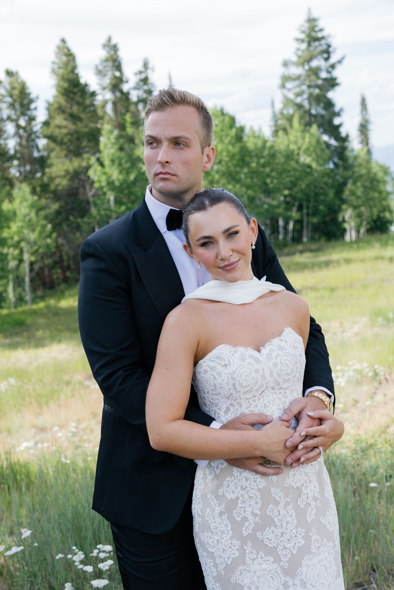 A romantic wedding couple embraces in a sun-lit meadow with evergreen trees in the background.