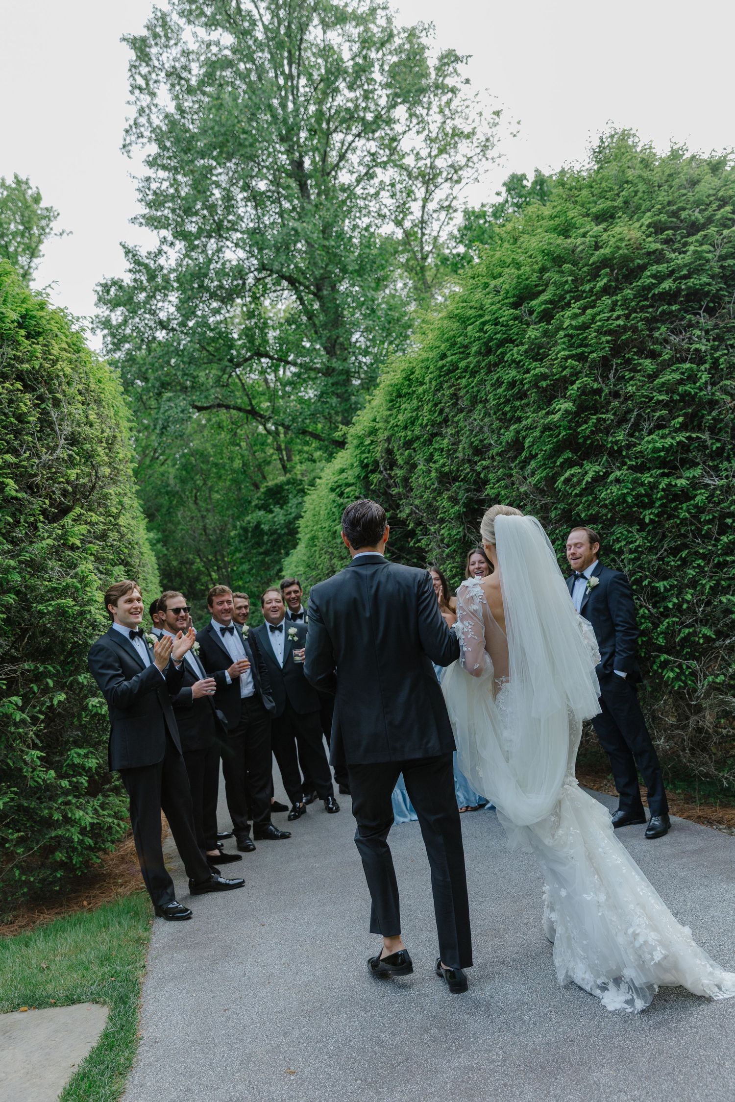 Wedding party gathered on a garden path surrounded by lush green hedges and trees during an outdoor ceremony.