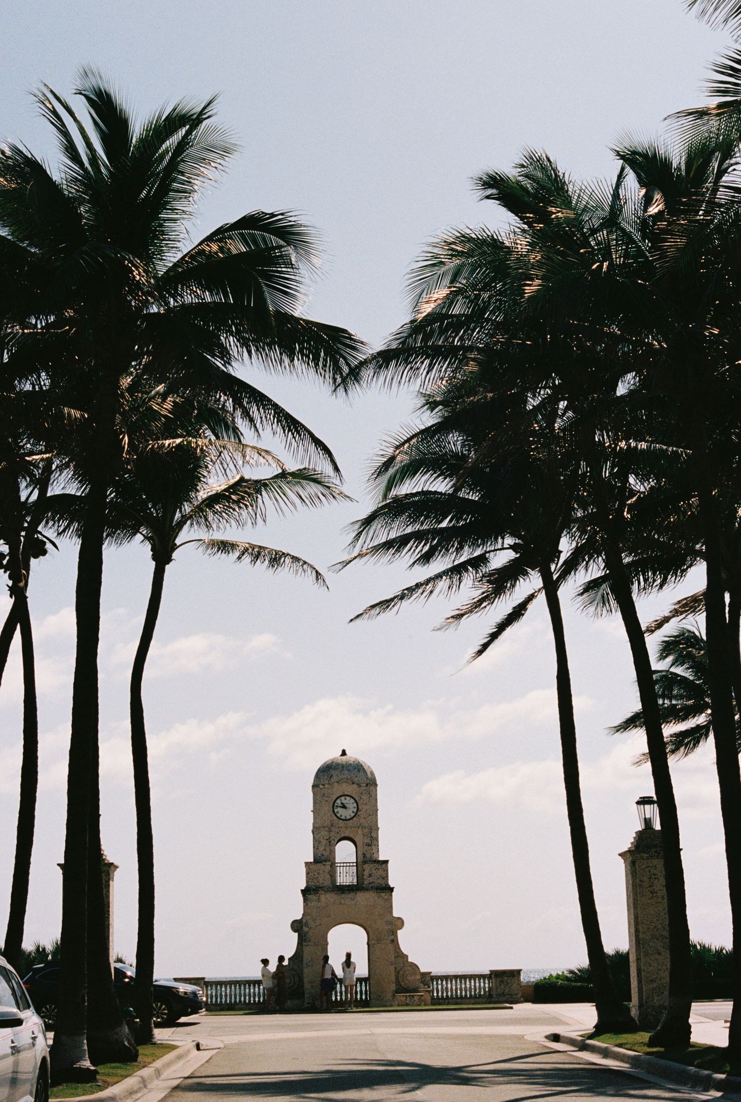 Palm trees frame the iconic Worth Avenue clock tower on a paved road in Palm Beach, Florida against a cloudy sky.