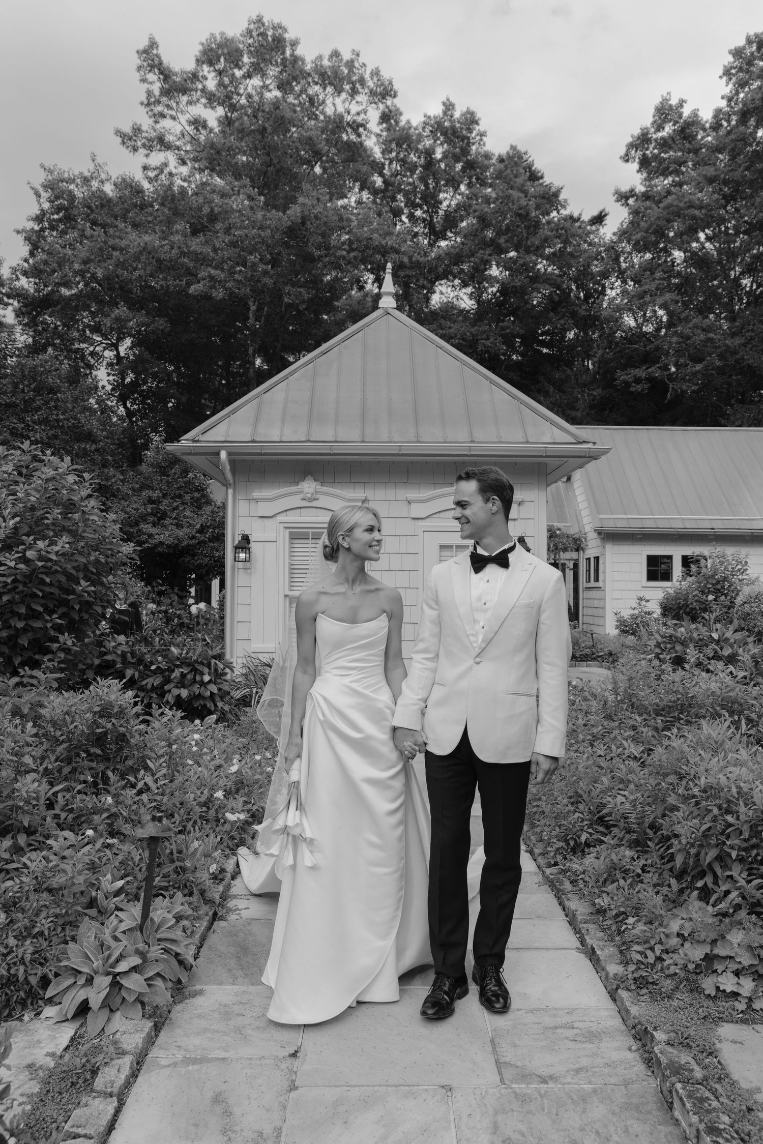 Black and white photo of a bride and groom holding hands and walking down a stone garden path past a small cottage.