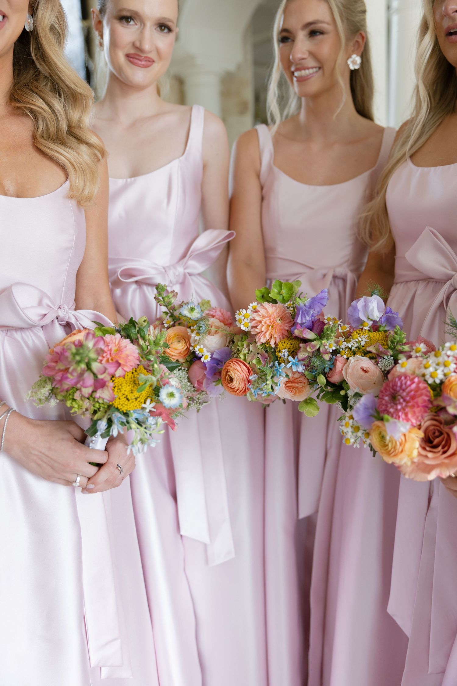 Bridesmaids in soft pink dresses stand together holding colorful spring bouquets with mixed flowers.