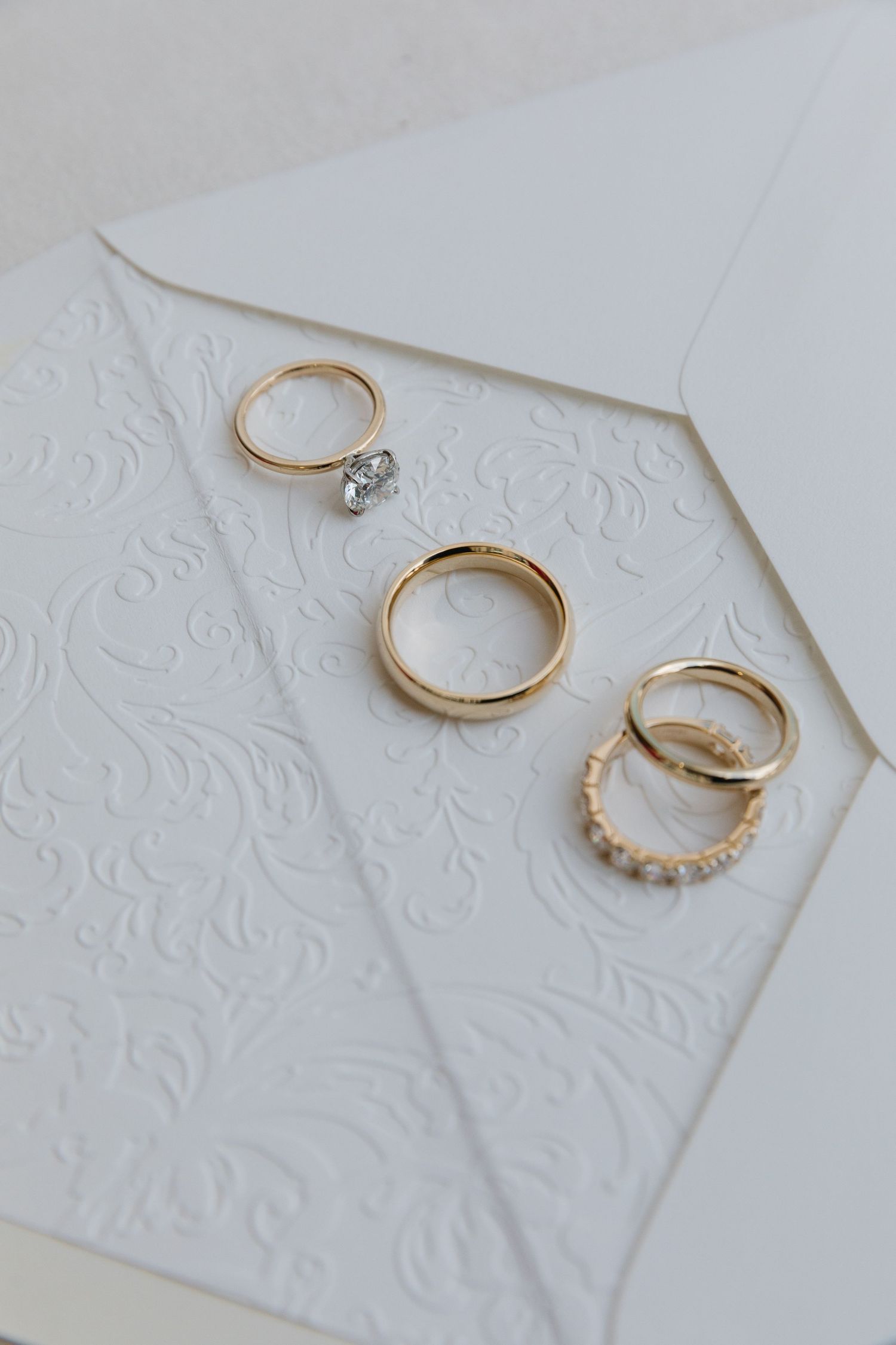 Delicate gold wedding rings and engagement bands arranged on white embossed paper backgrounds in a series of photos.