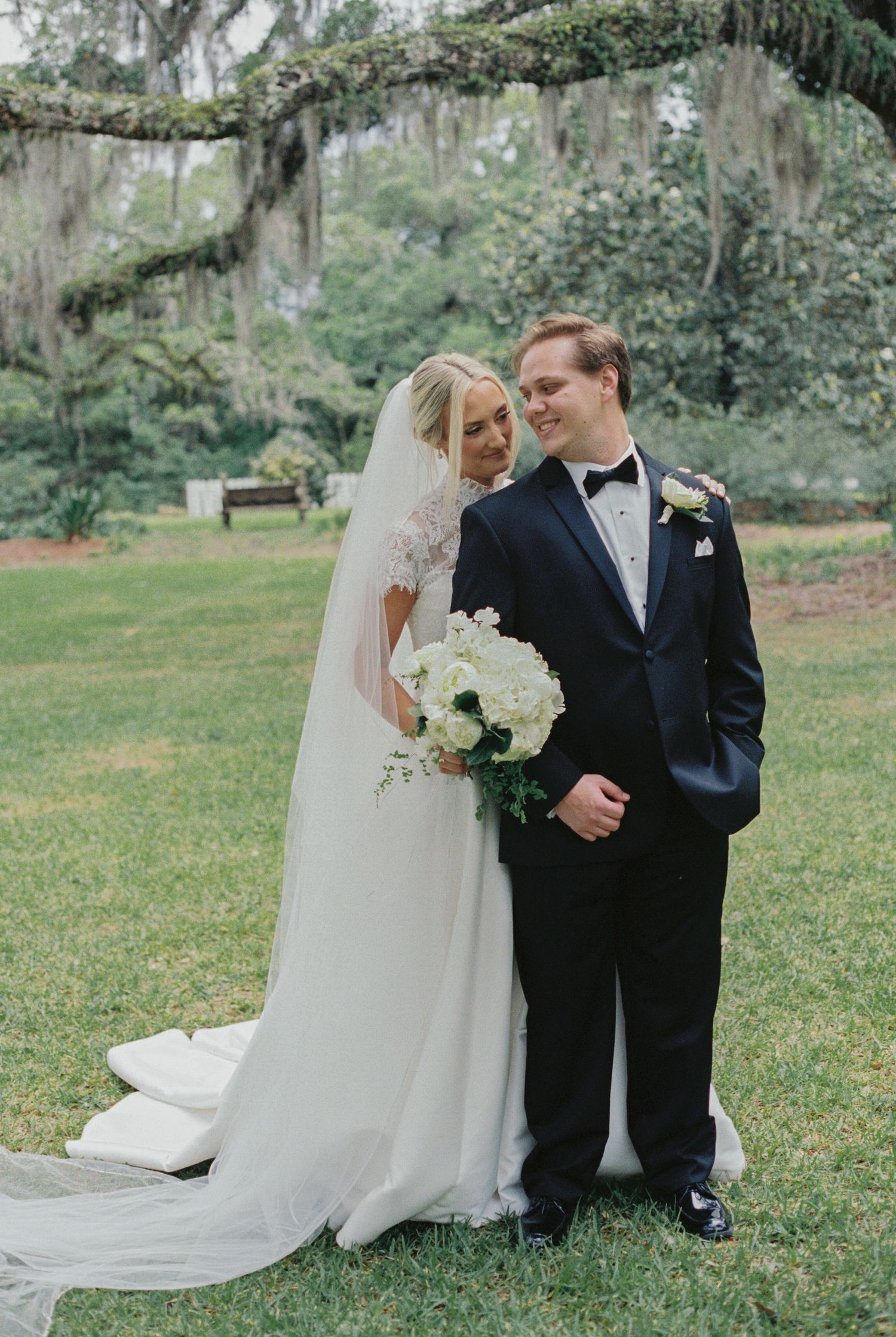 Bride in lace gown with veil holds white bouquet beside groom in navy tuxedo under Spanish moss trees.