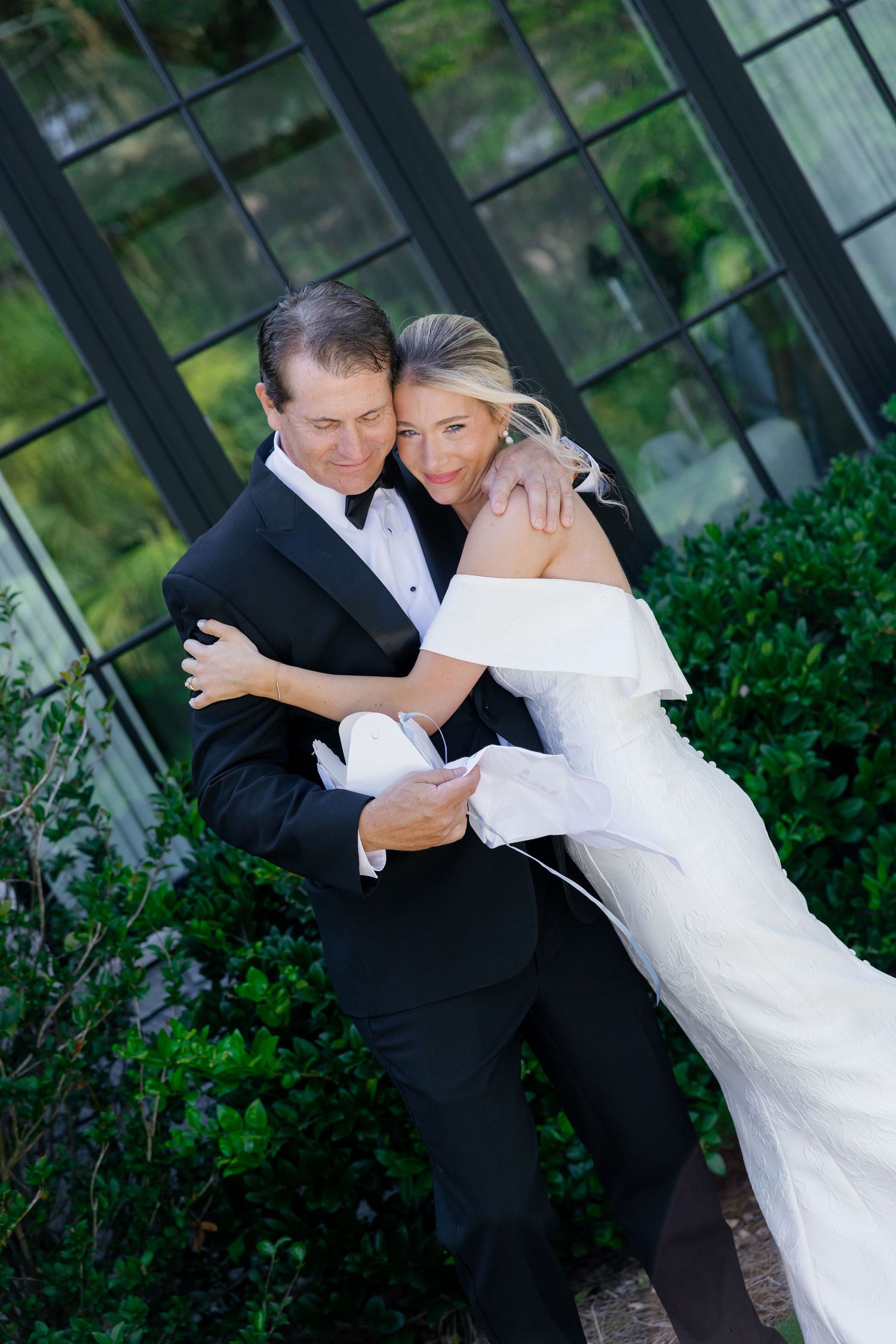 A couple in formal wedding attire dance together in an outdoor garden setting with greenery and black railings.