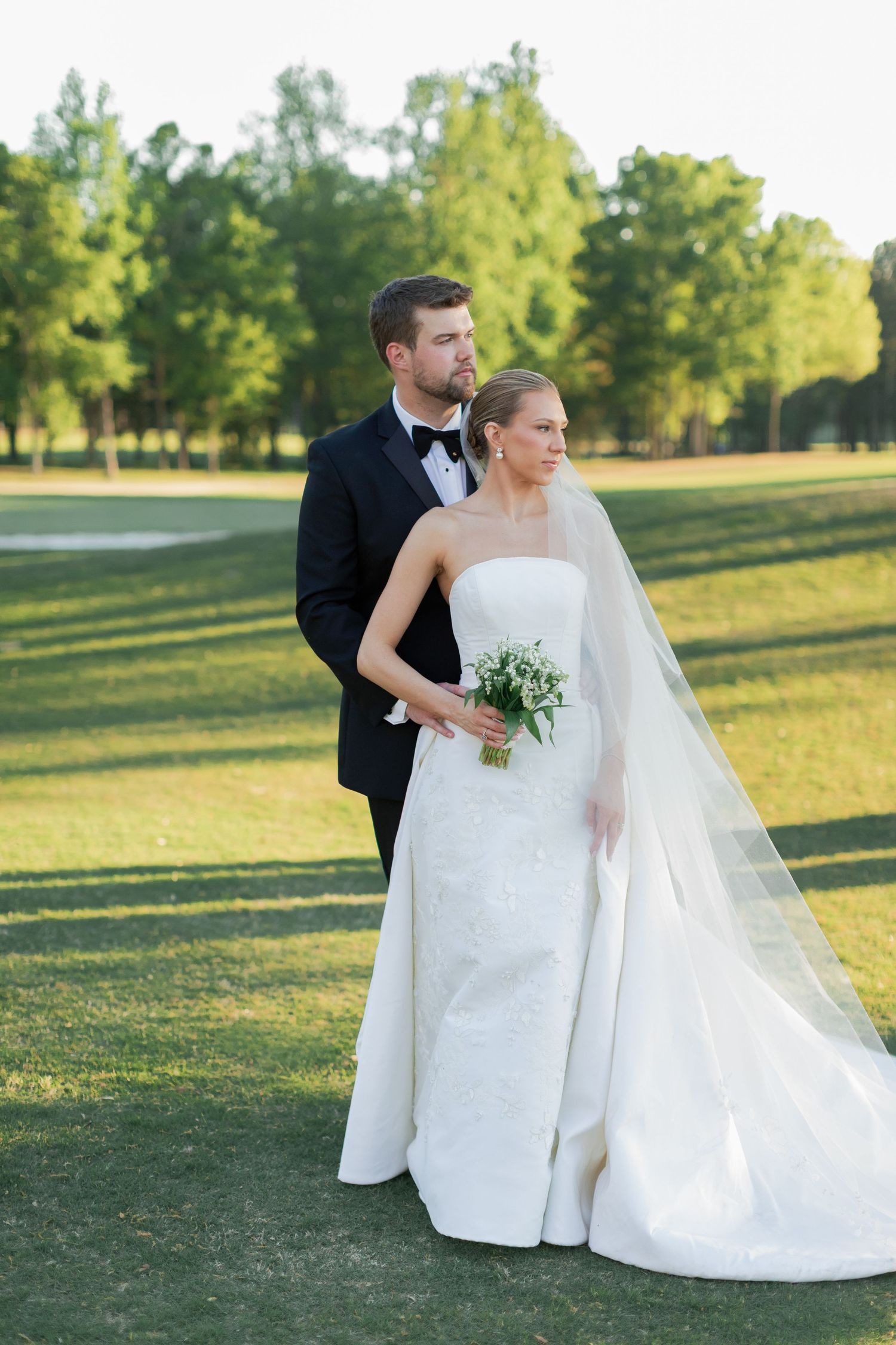 Bride in strapless white gown with veil holds bouquet as groom in black tuxedo embraces her on lush green golf course.