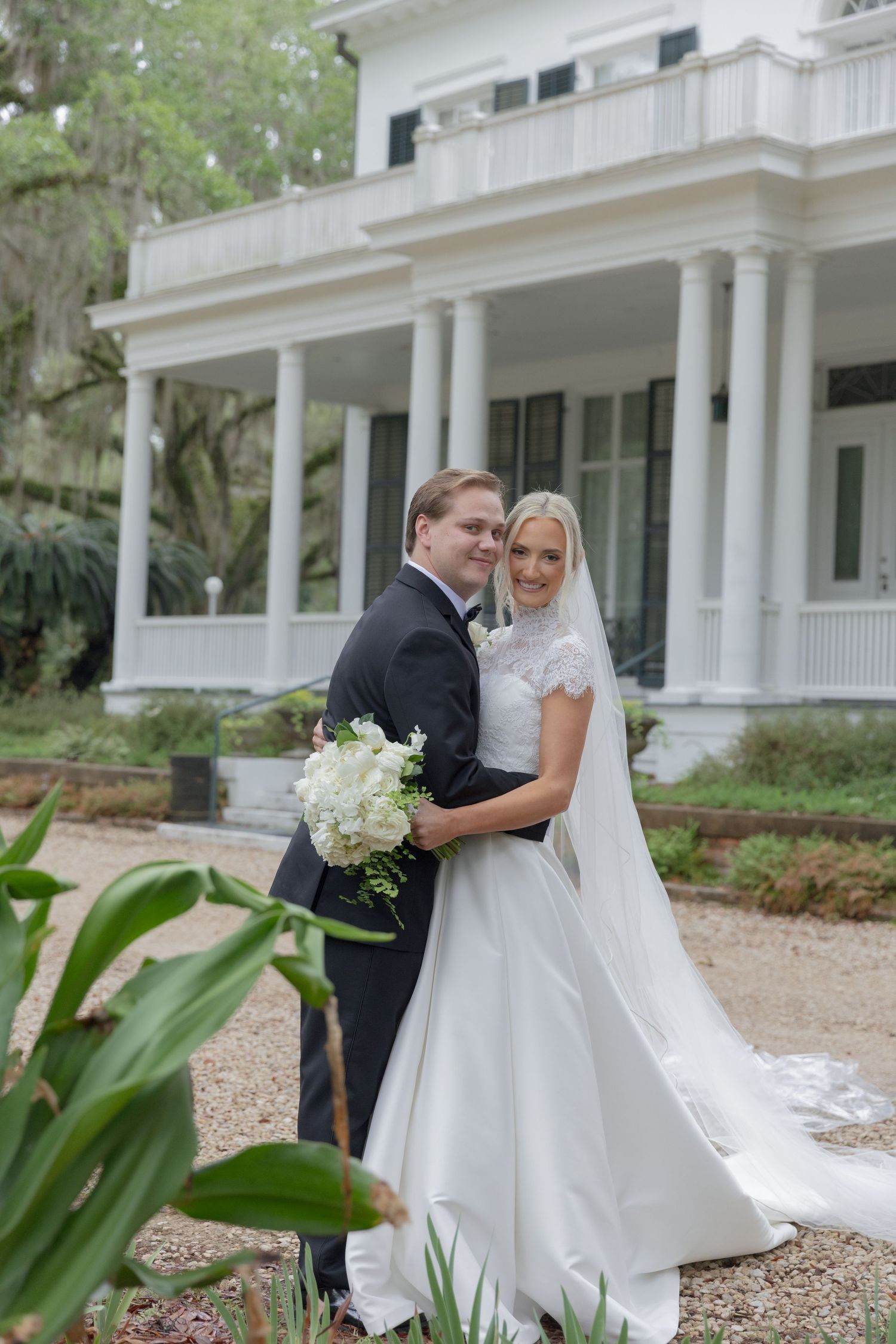 Bride and groom pose together in front of a white Southern mansion, bride holding white bouquet in elegant wedding attire.