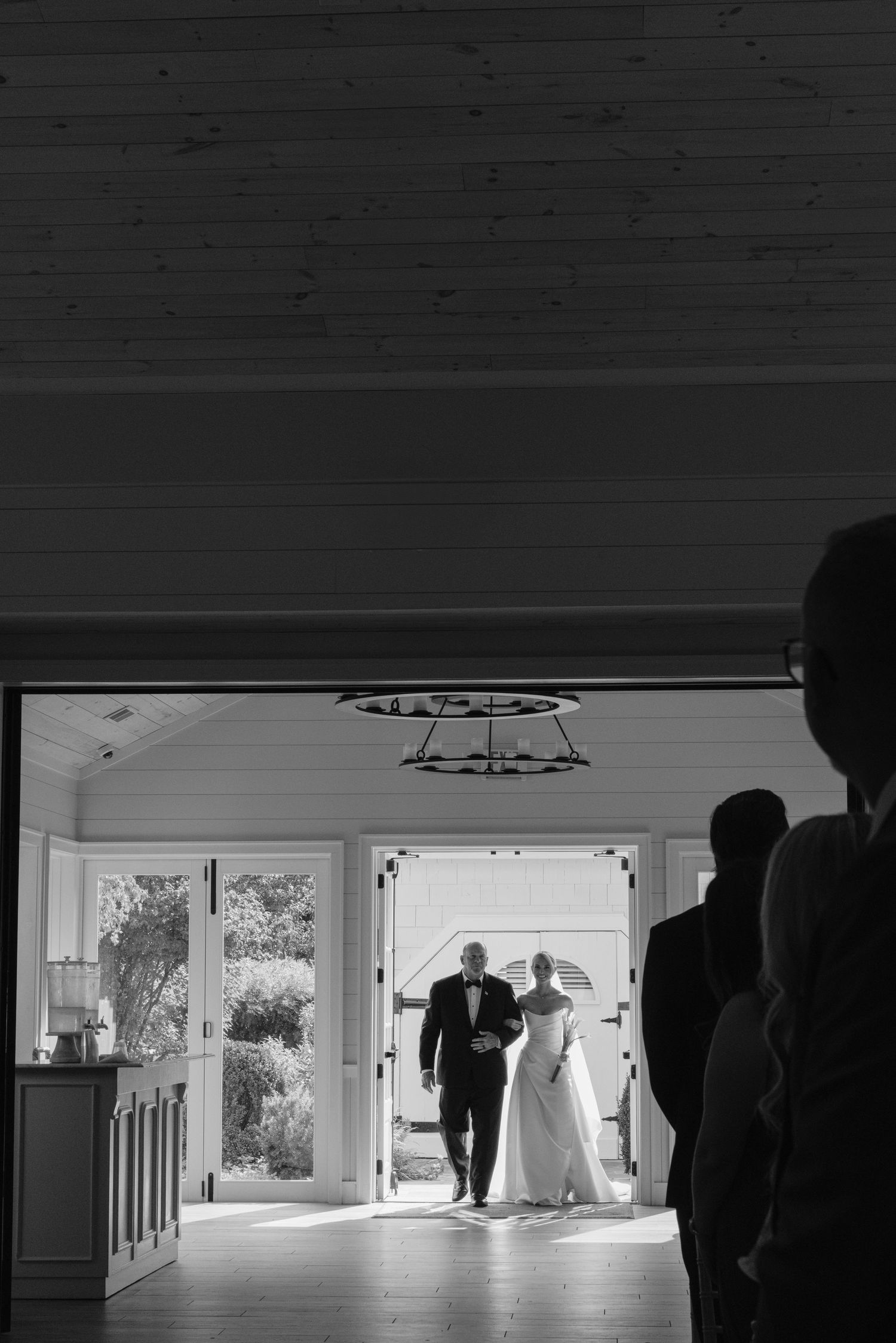 Black and white photo of a father walking a bride in a wedding gown down the aisle as guests watch from the foreground.