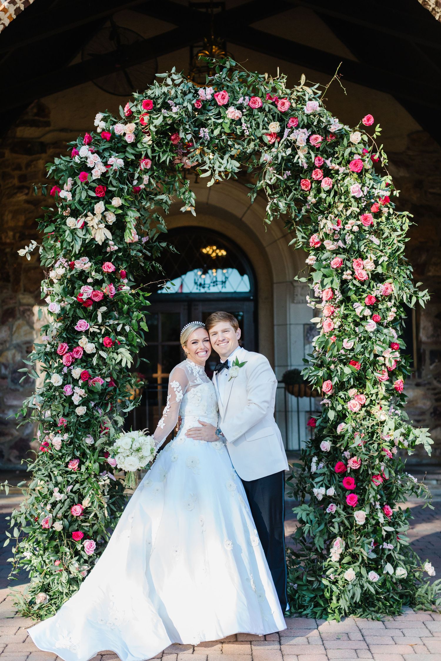 Romantic wedding couple poses under floral archways decorated with pink roses and greenery at a stone venue.