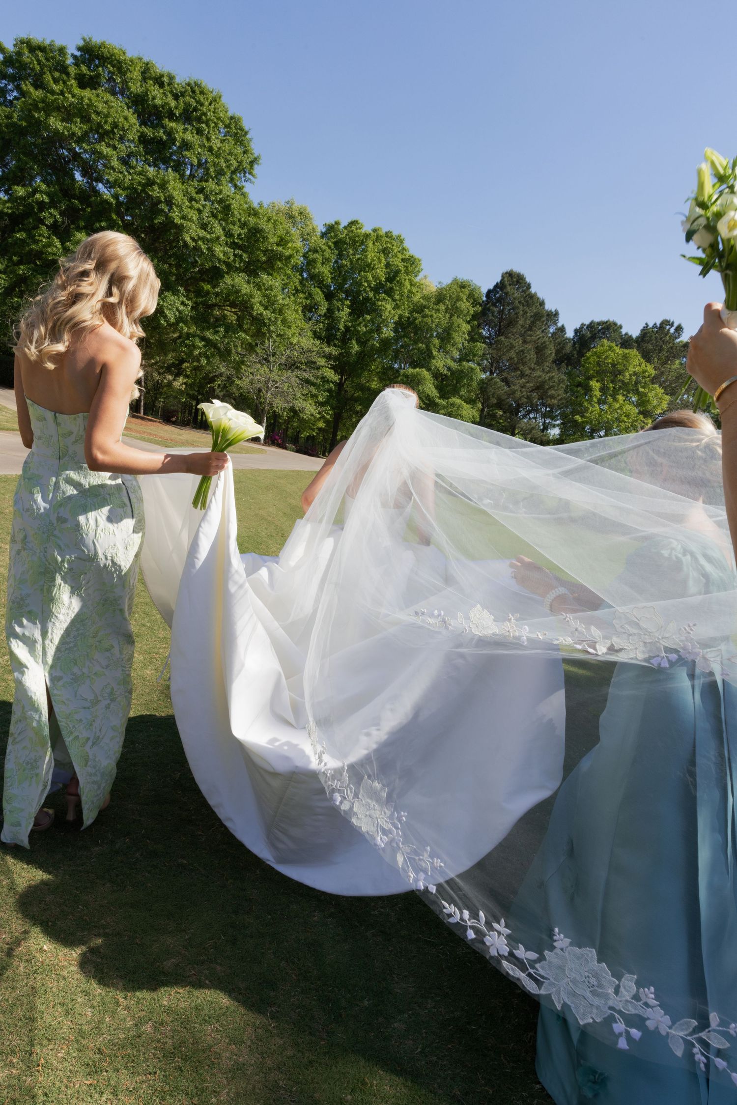 Bridesmaids in sage green gowns help arrange a bride's white lace wedding dress train on a sunny outdoor lawn.