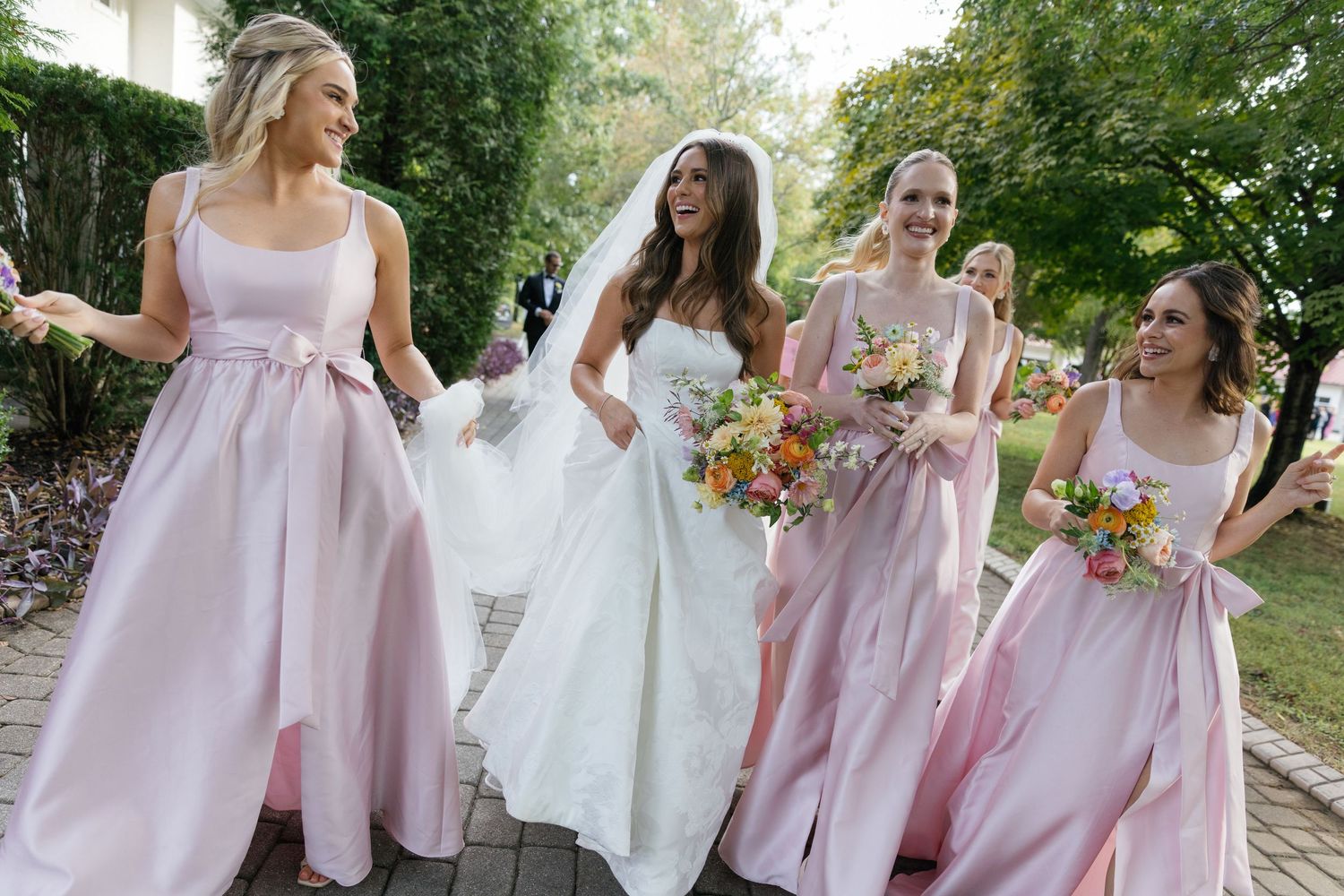 Wedding party wearing pink and white dresses poses together outdoors in a garden setting.
