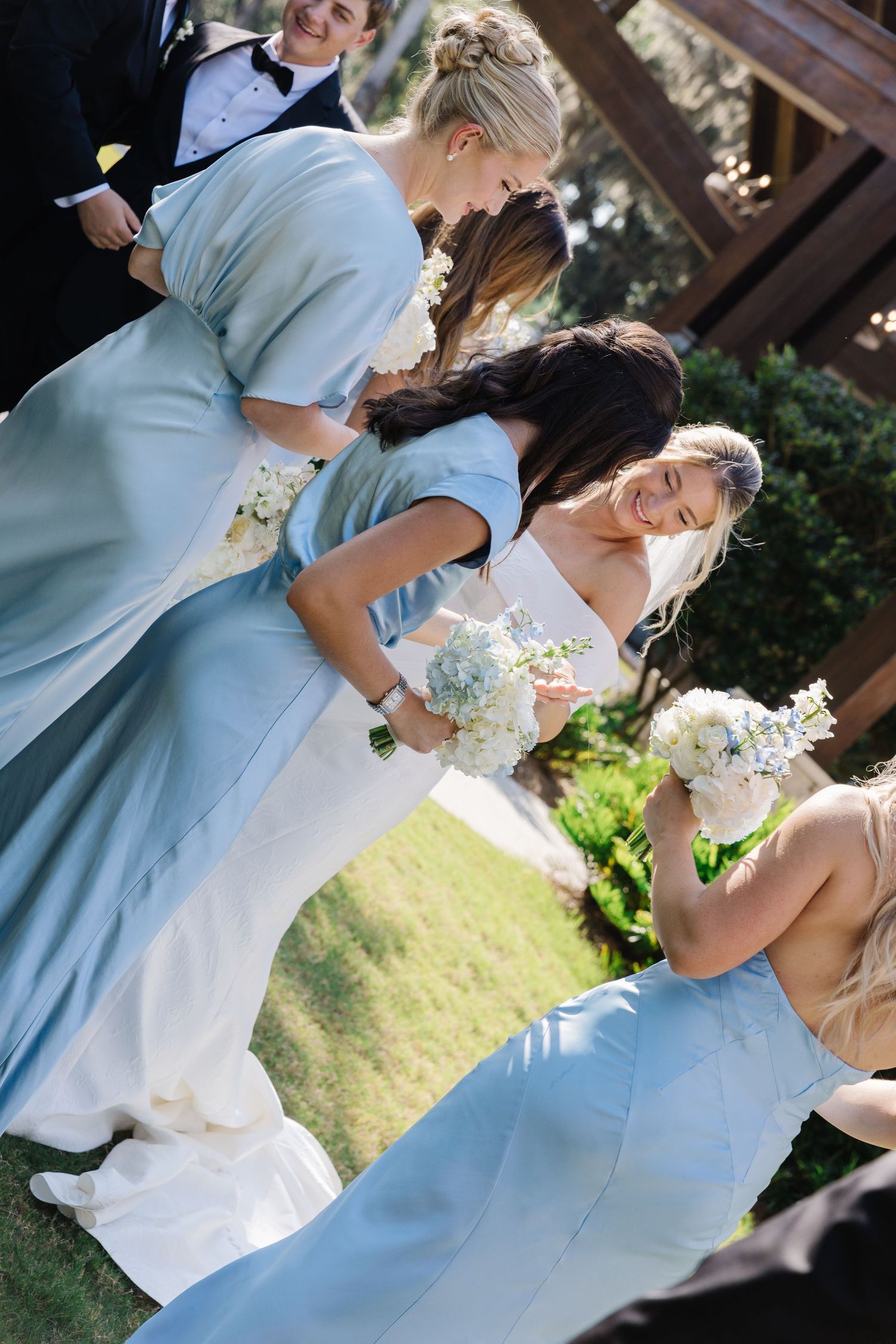 A romantic wedding couple embraces and kisses in a garden setting with bridesmaids in light blue dresses nearby.