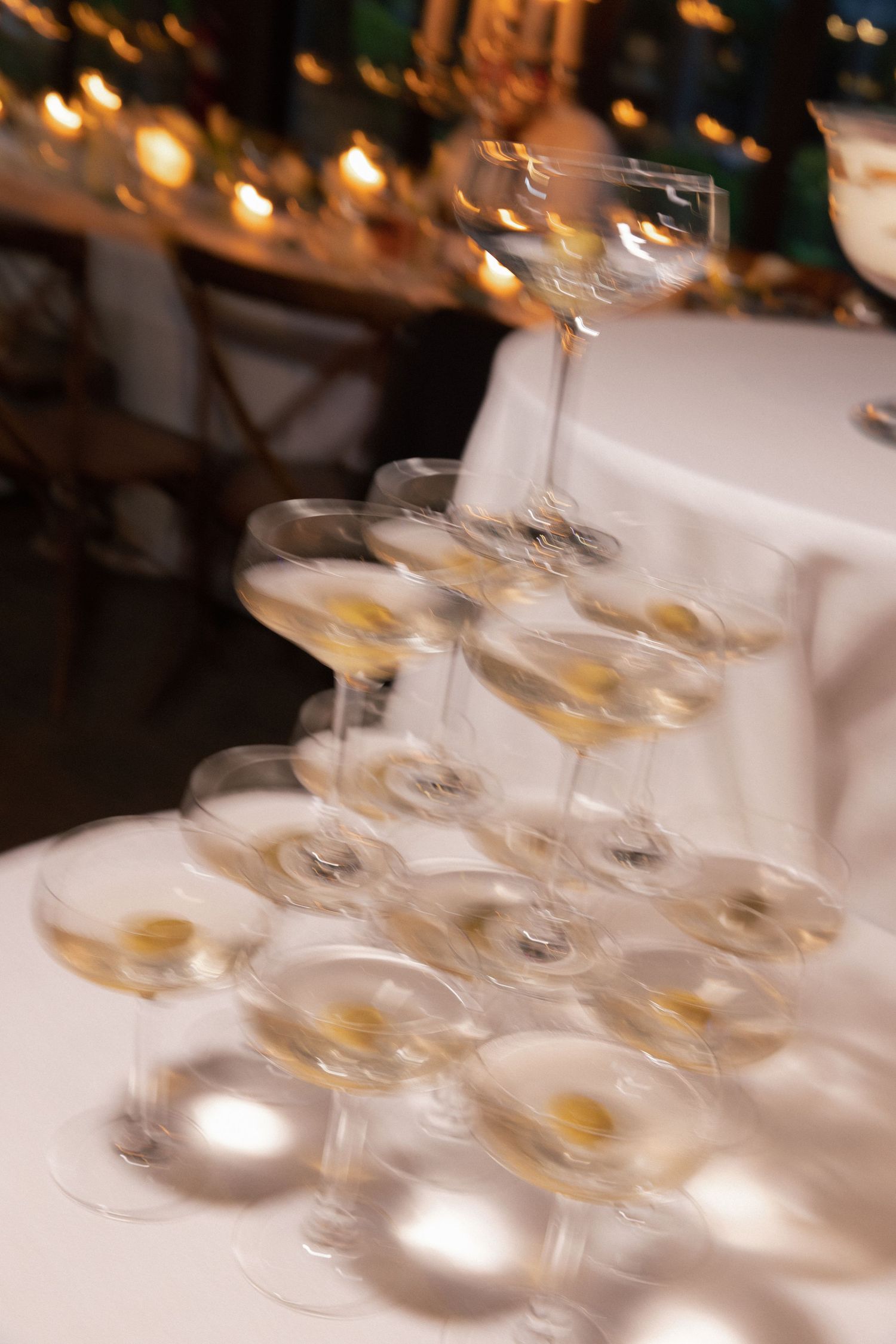 A blurred tower of champagne coupes filled with golden martinis stacked on a white tablecloth at a candlelit wedding reception.