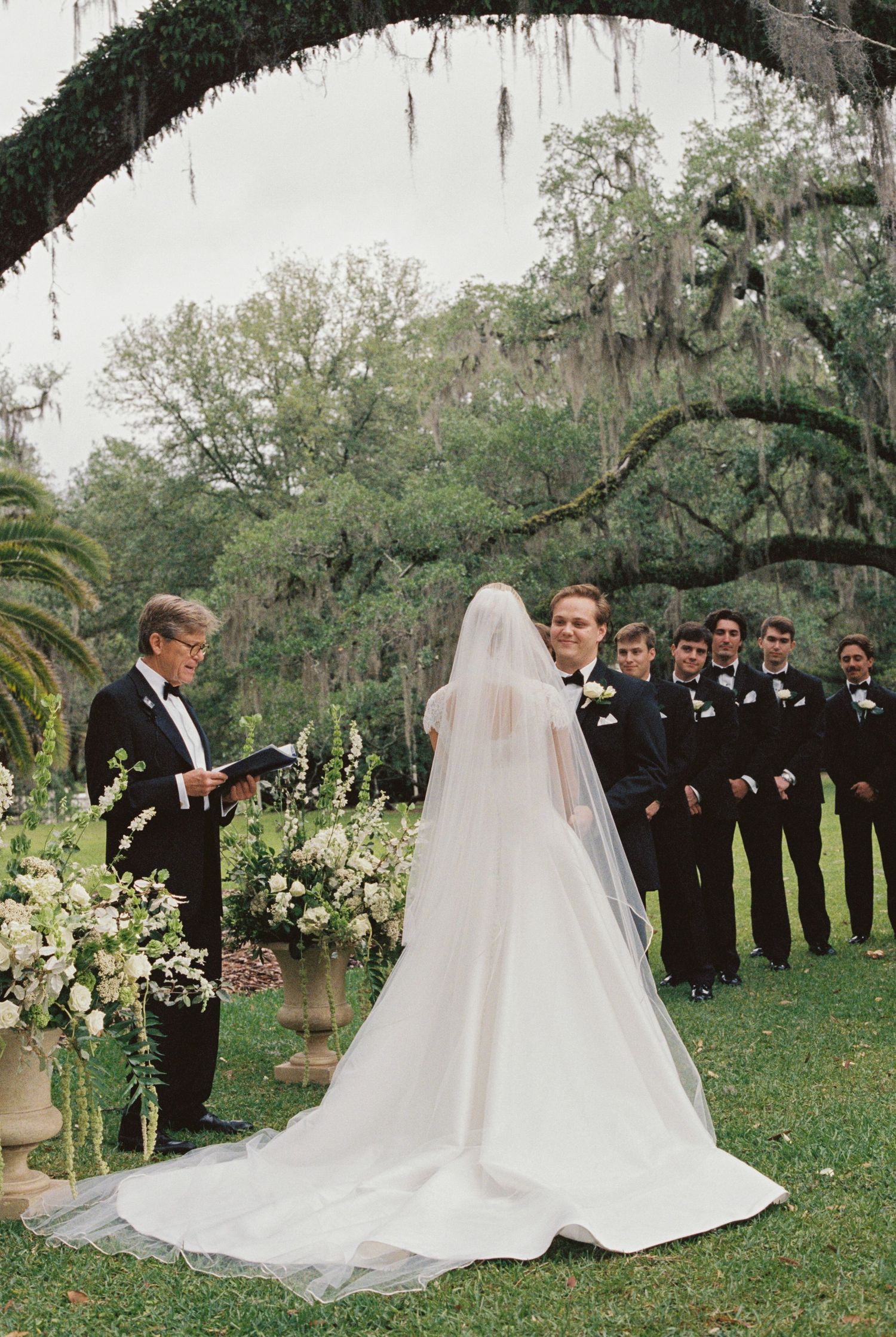Outdoor wedding ceremony under Spanish moss-draped oak trees, bride in white gown with veil, groom and groomsmen in black tuxedos.