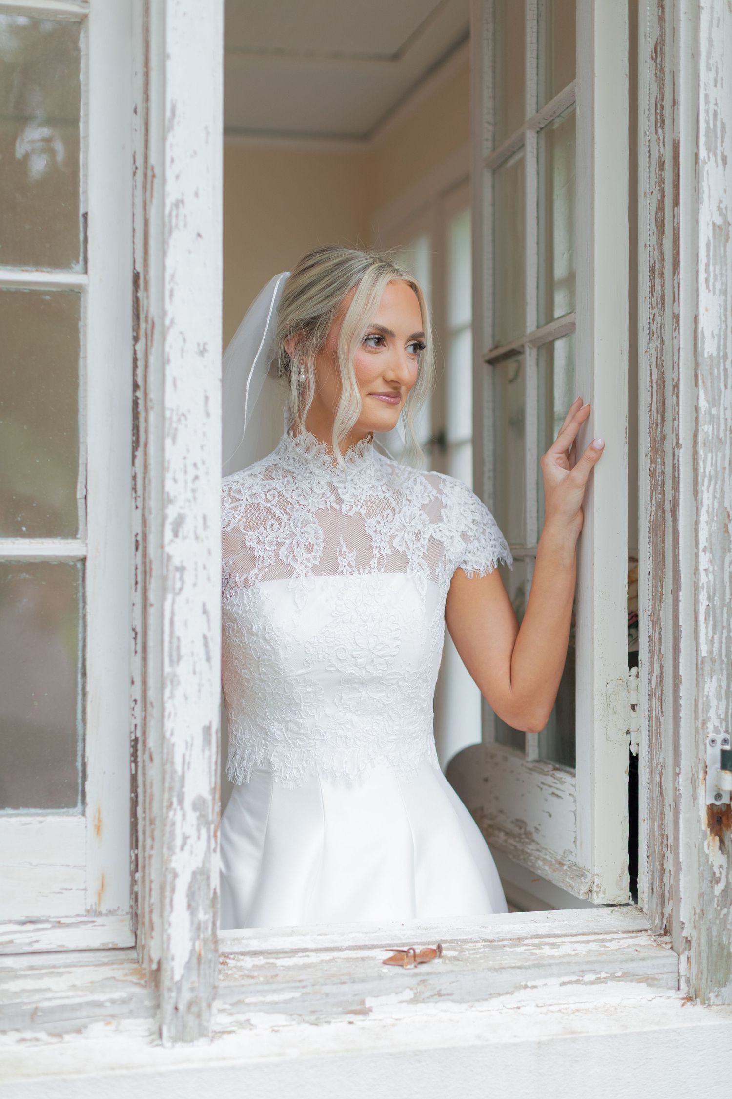 Elegant bride in lace high-neck wedding dress with veil standing in vintage white doorway, smiling softly.