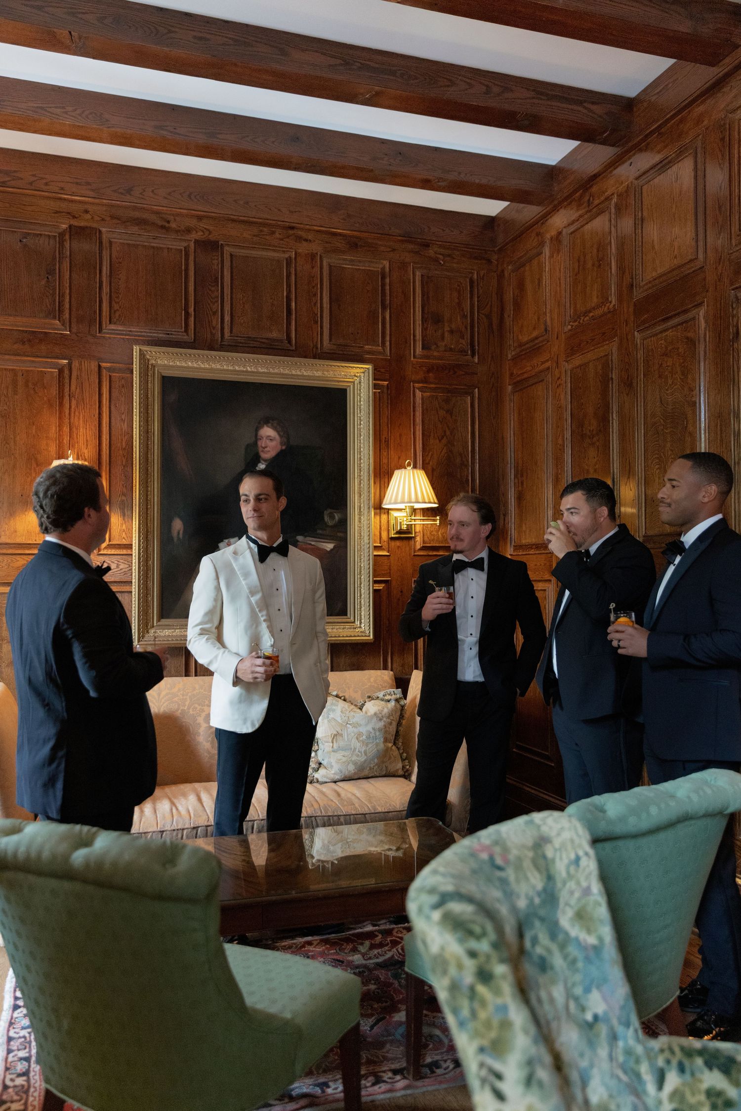 Groomsmen in tuxedos and a white dinner jacket chat in a wood-paneled room with a portrait and green armchairs.