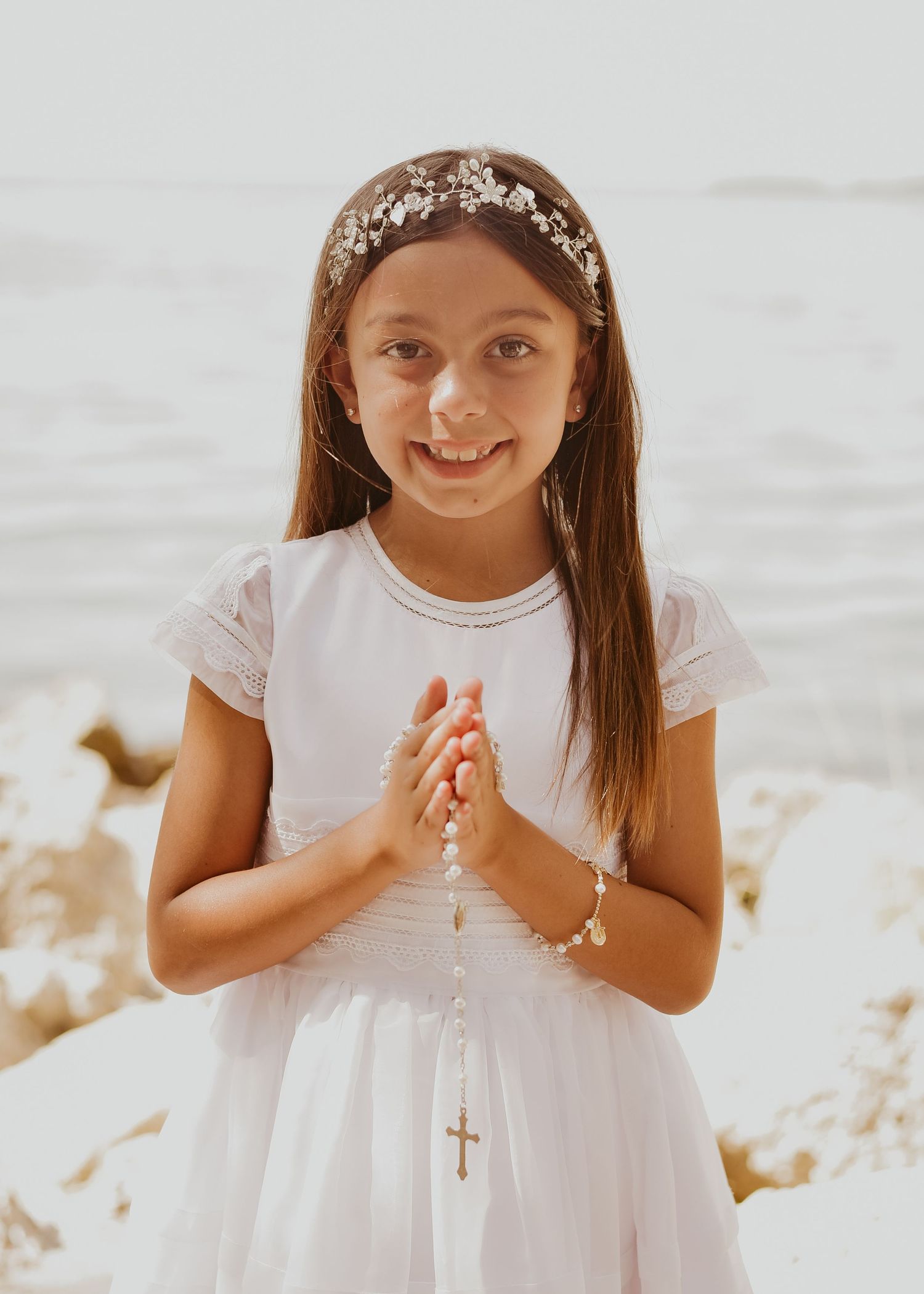Child in white dress by beach holding rosary during First Communion portrait session