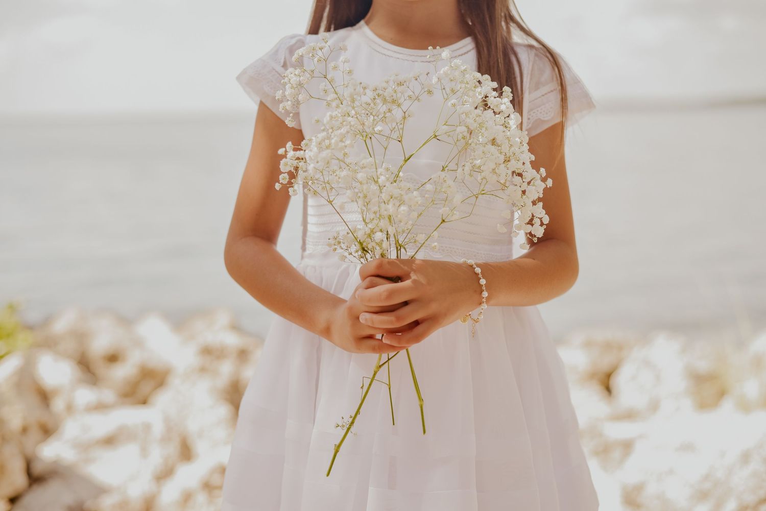 Child in white dress by beach holding flowers during First Communion portrait session