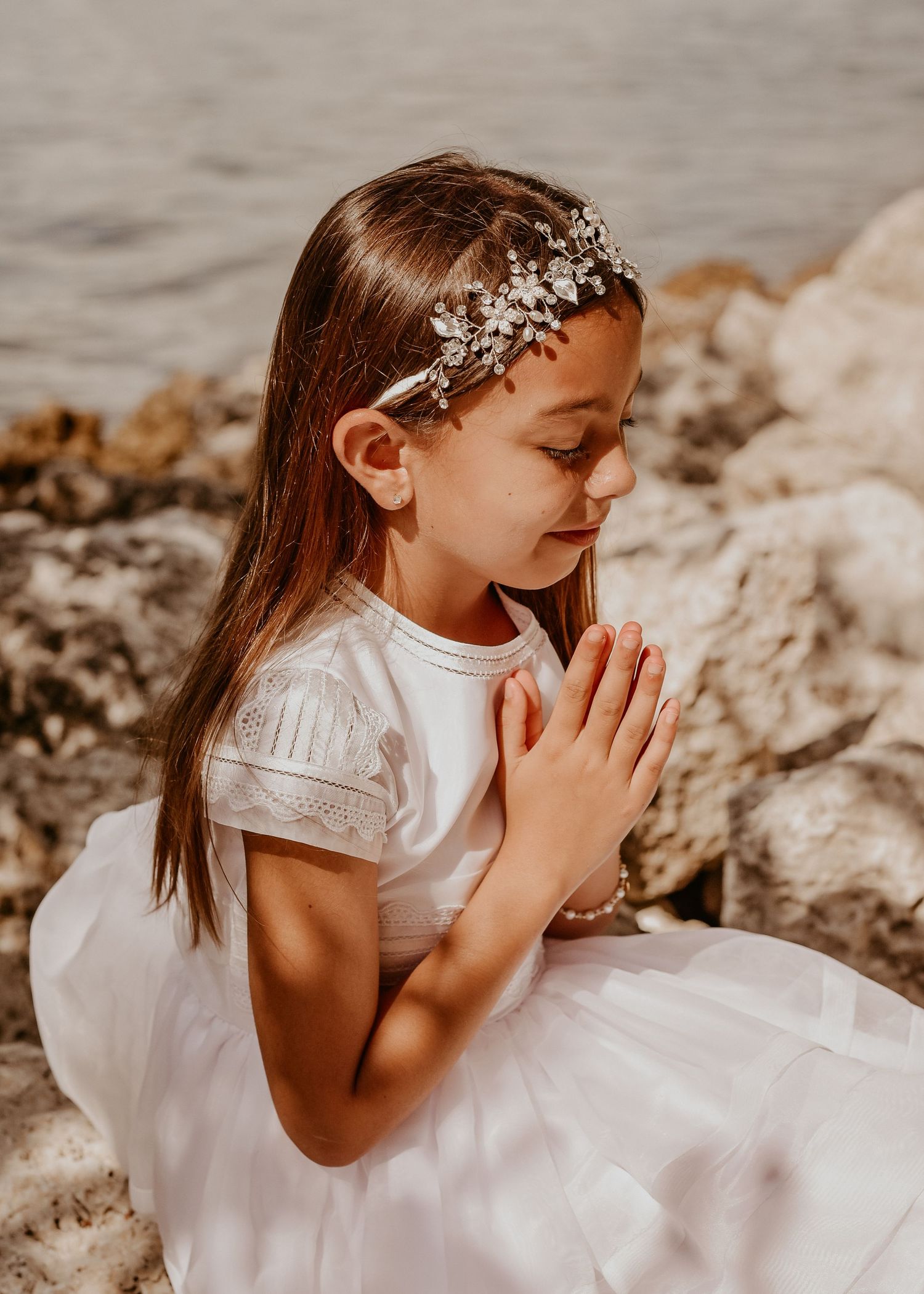 Child in white dress by beach holding rosary during First Communion portrait session