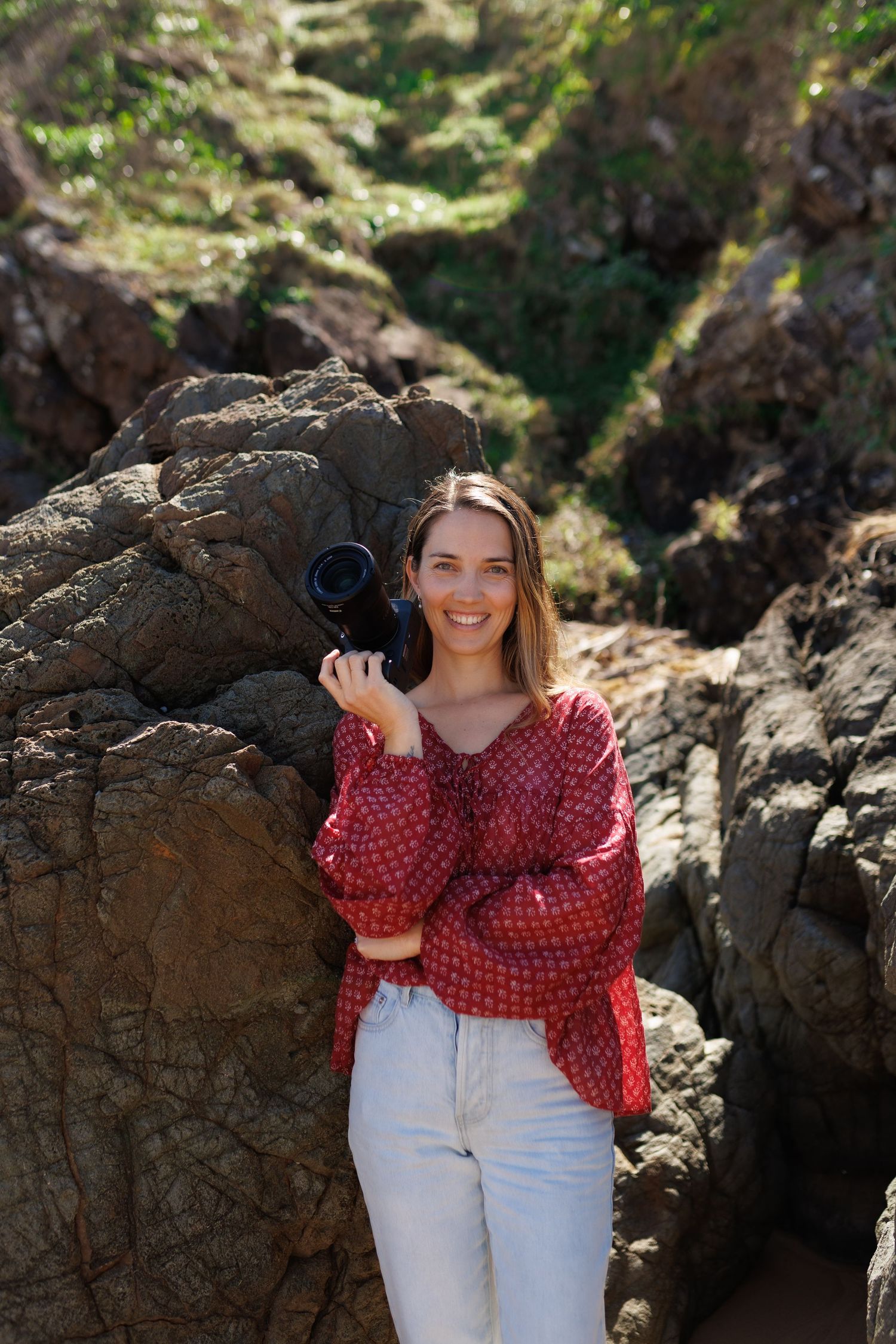 Woman in red polka dot top and white pants poses against rocky coastal backdrop.