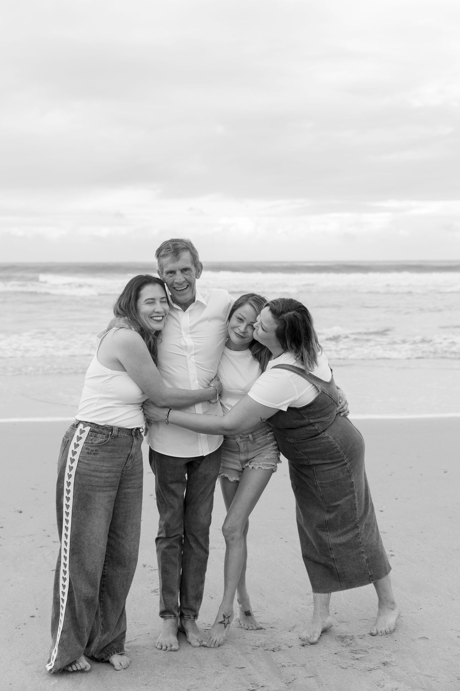 Black and white photo of a happy family of four hugging and laughing together on a sandy beach. Coffs Coast photographer.
