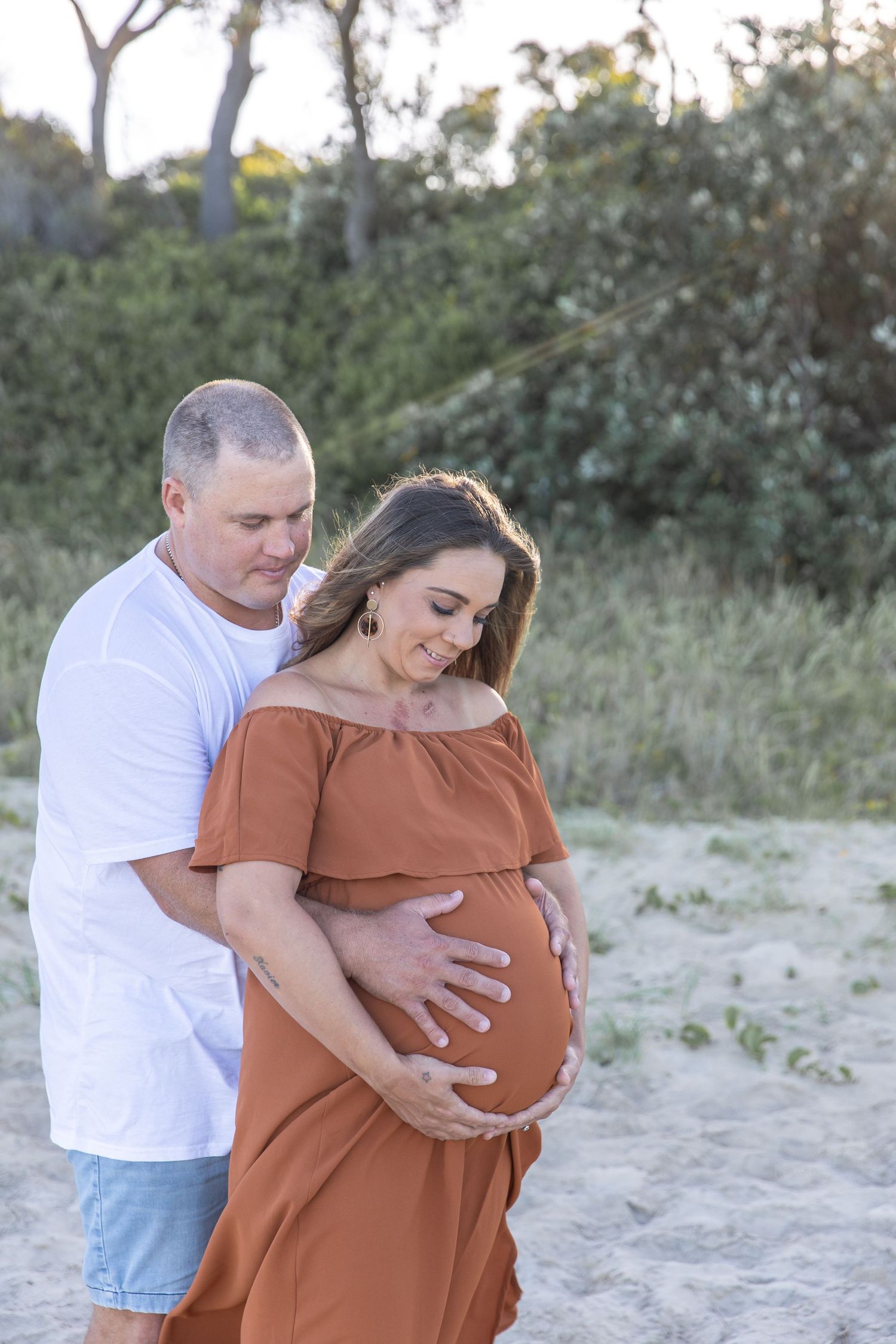 Couple sharing a tender moment on sandy beach, man embracing pregnant woman in rust off-shoulder dress, both looking at baby bump.