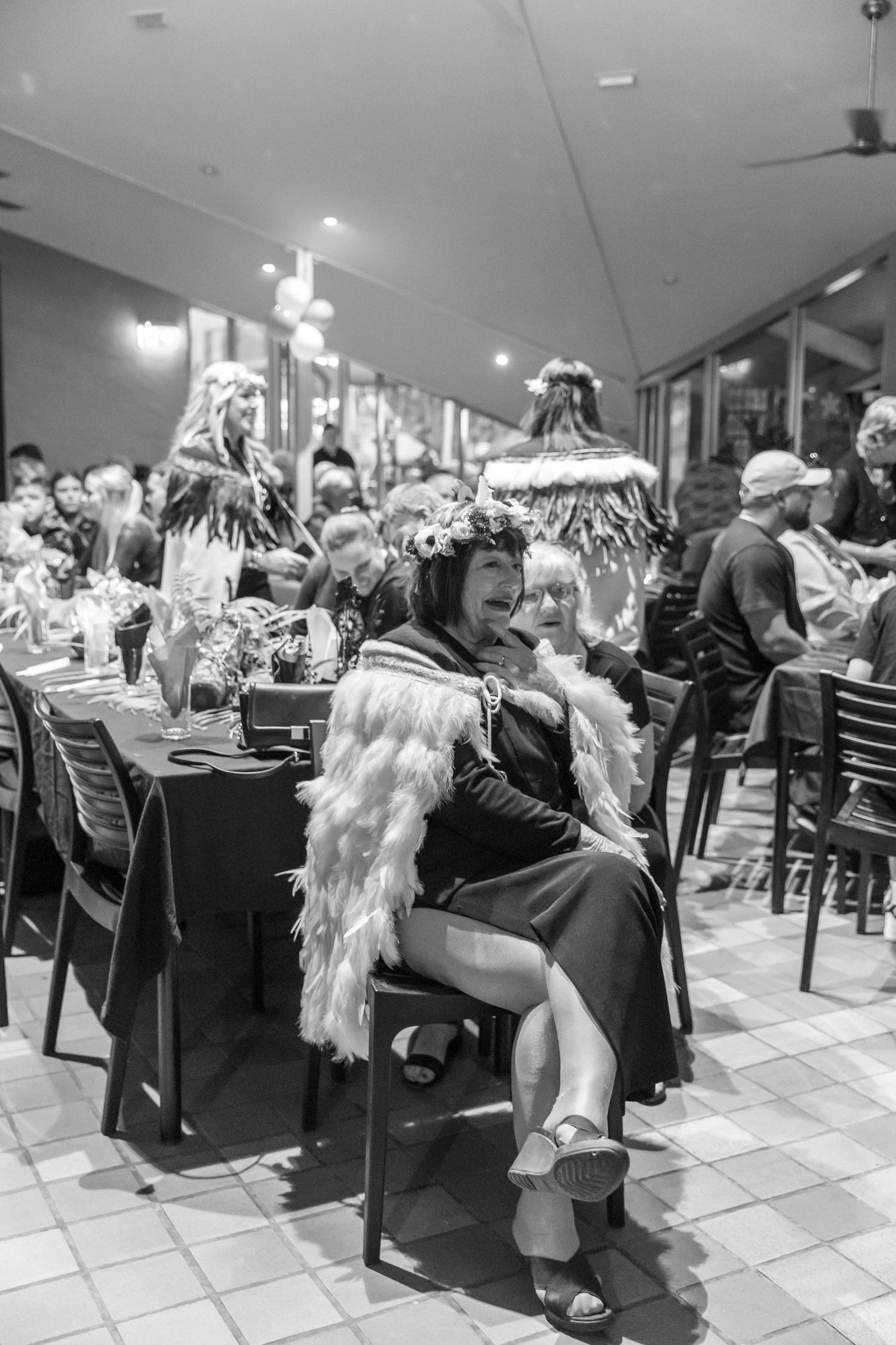 Black and white photo of a woman in feathered wrap and floral crown seated at a table during a formal event. Coffs Coast photographer.