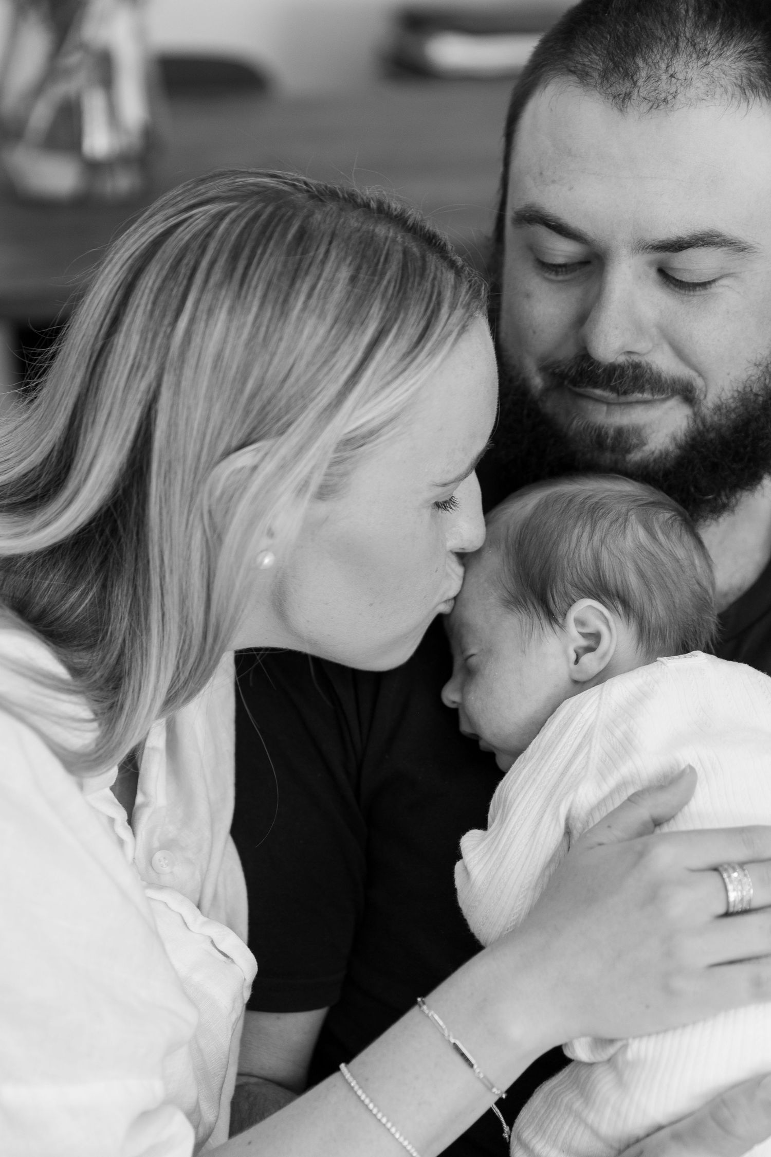 Black and white photo of parents lovingly kissing and gazing at their newborn baby wrapped in white.