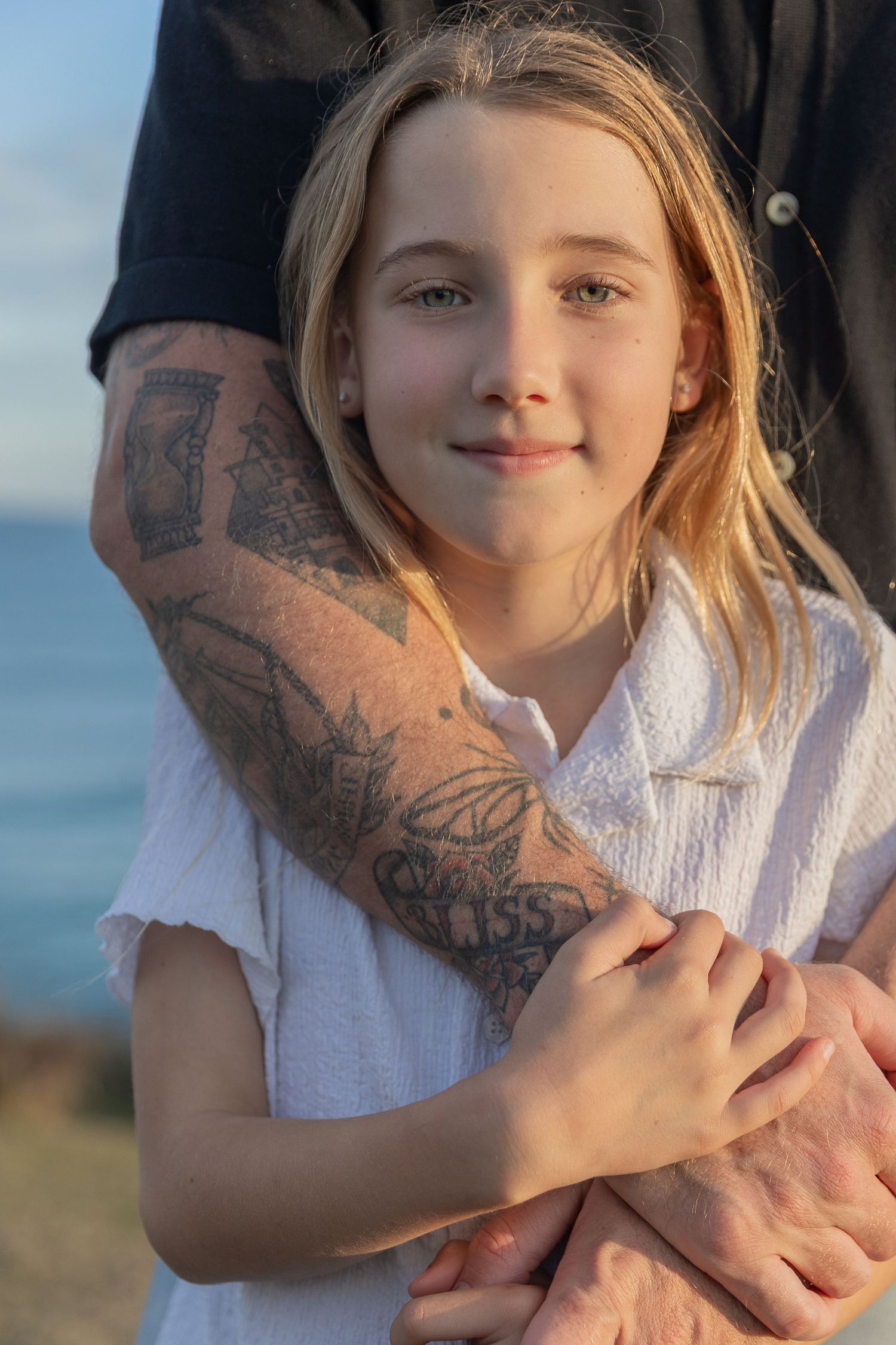 Close up portrait of a child and tattooed arm against a coastal background at sunset.