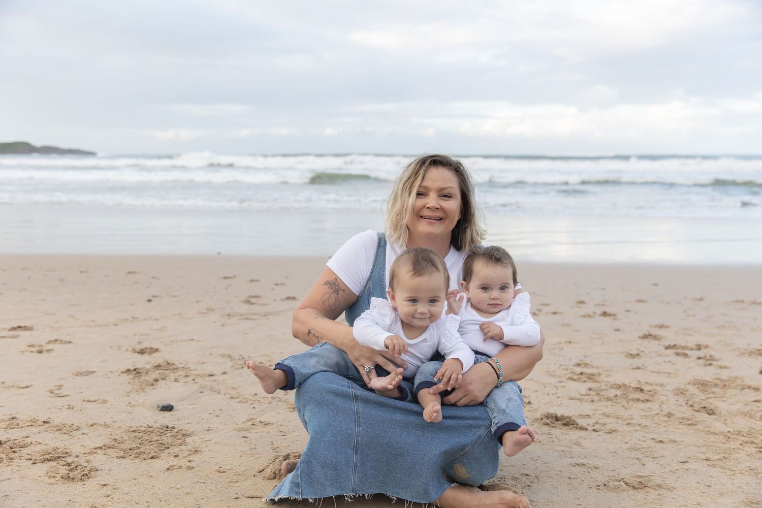 A woman sits on a sandy beach smiling with two young toddlers in her lap, ocean waves in the background.