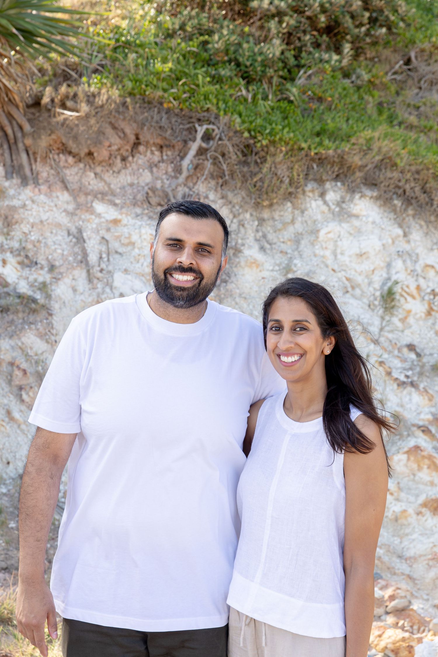 A smiling couple in matching white tops posing outdoors in front of a rocky cliff with green vegetation. Coffs Coast photographer.