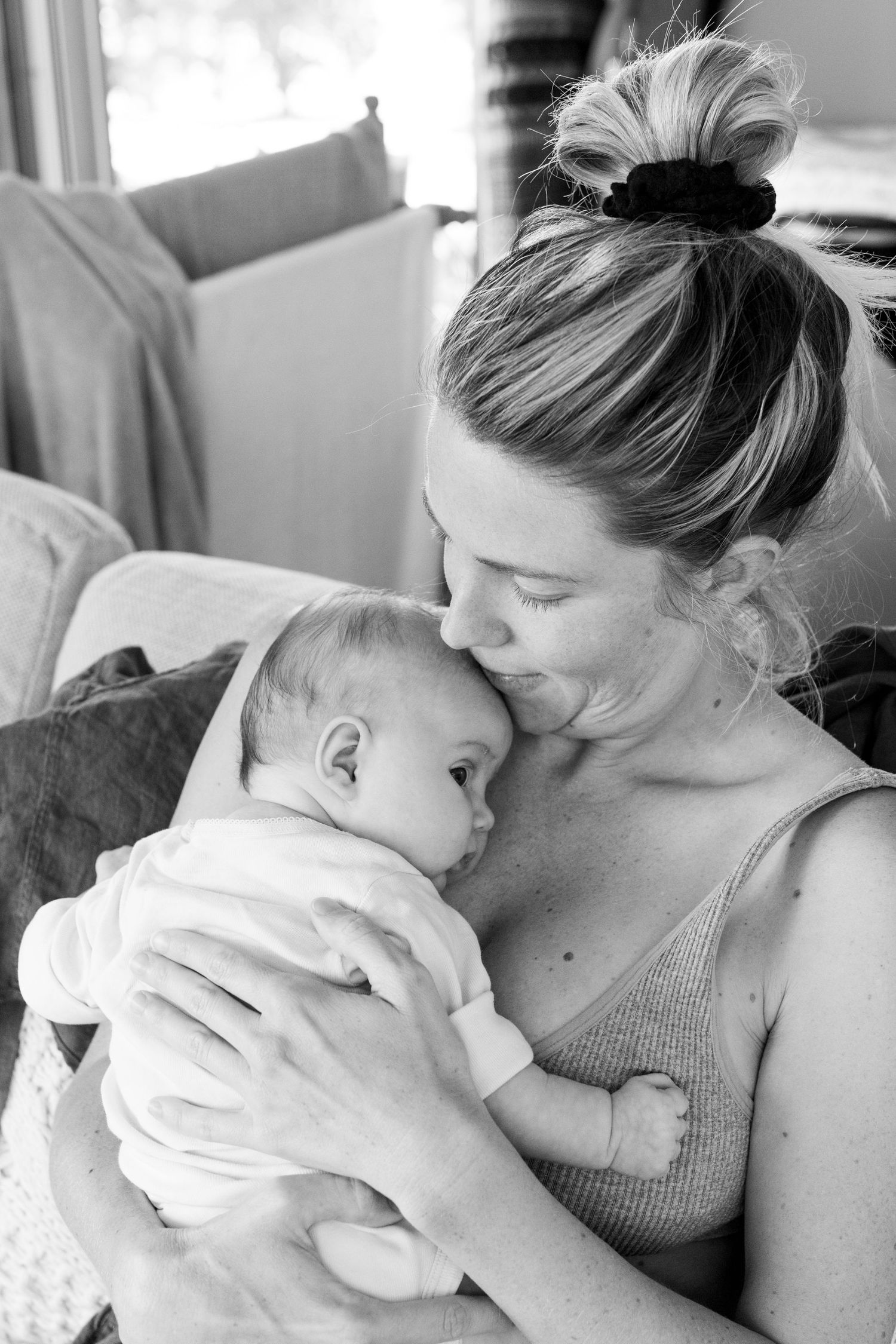 Black and white photo of a mother tenderly kissing her newborn baby while seated on a couch.