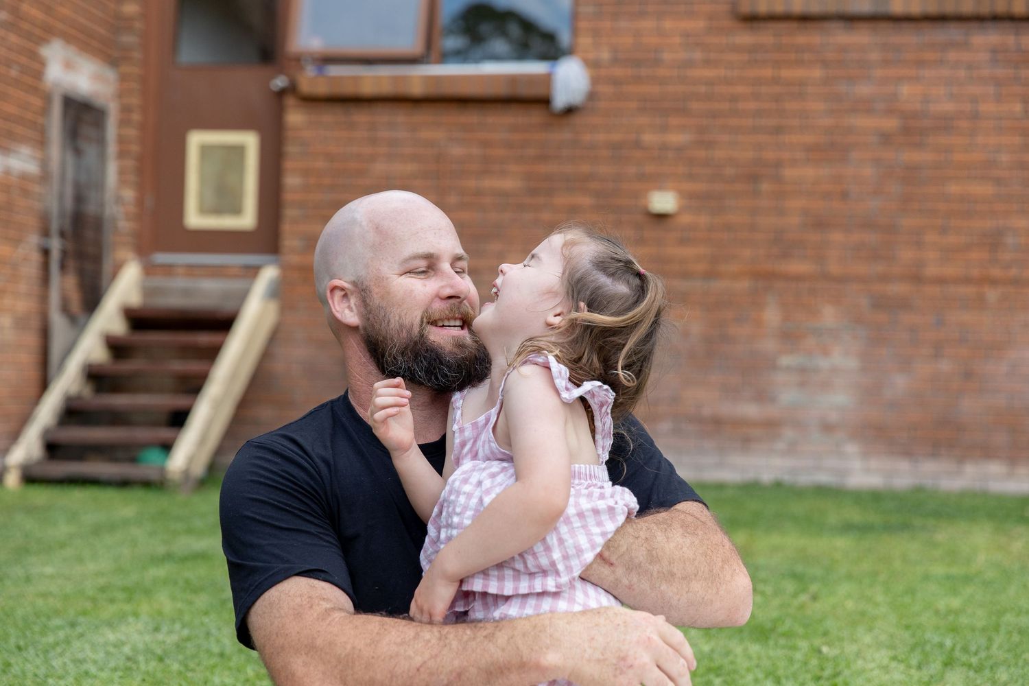 Bald bearded father holding and kissing his young daughter outdoors in backyard of brick home.