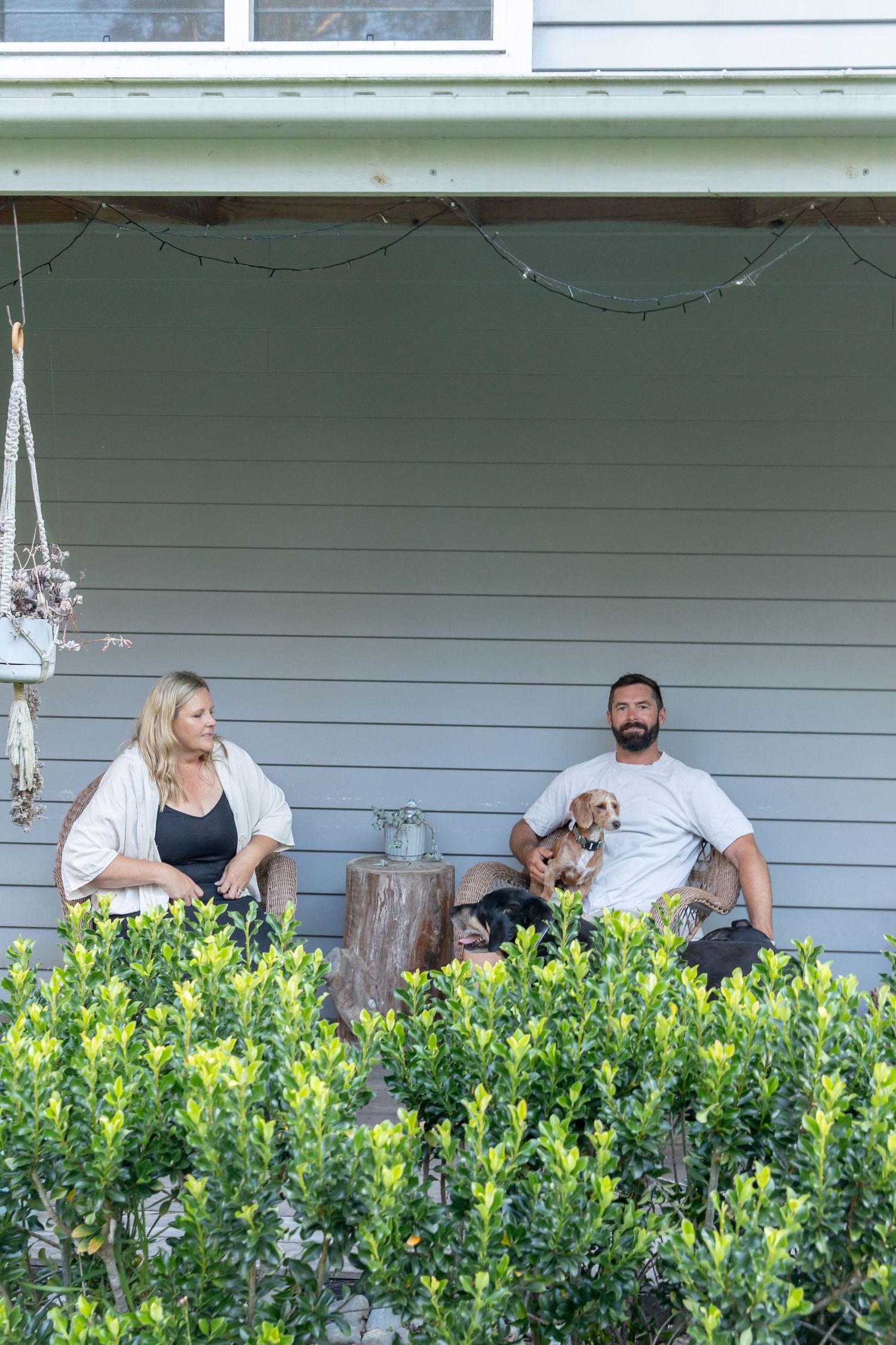 Family sits behind green bushes on their porch with their pet dog.