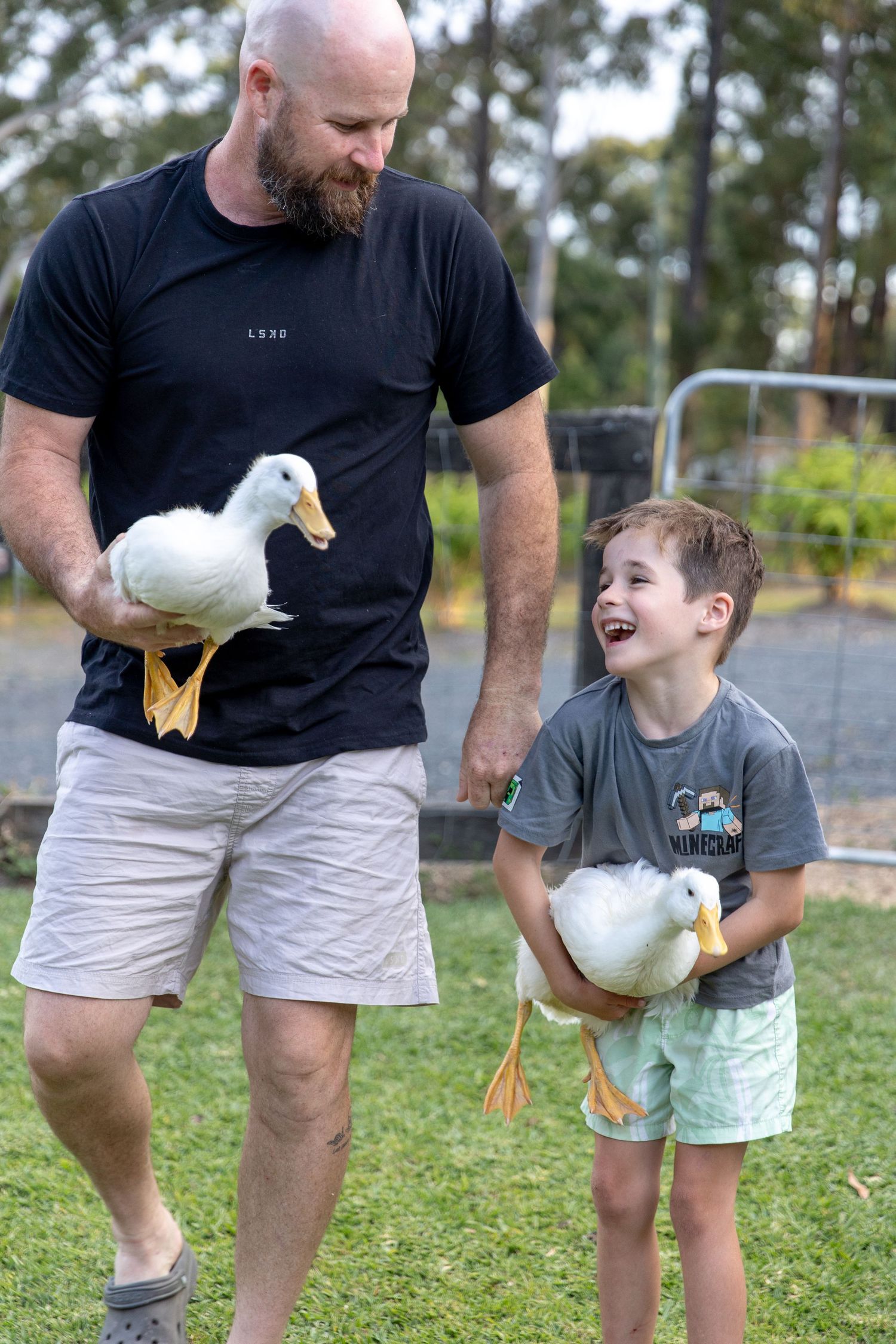 Parent and child in casual wear interacting with white duck on grassy lawn.