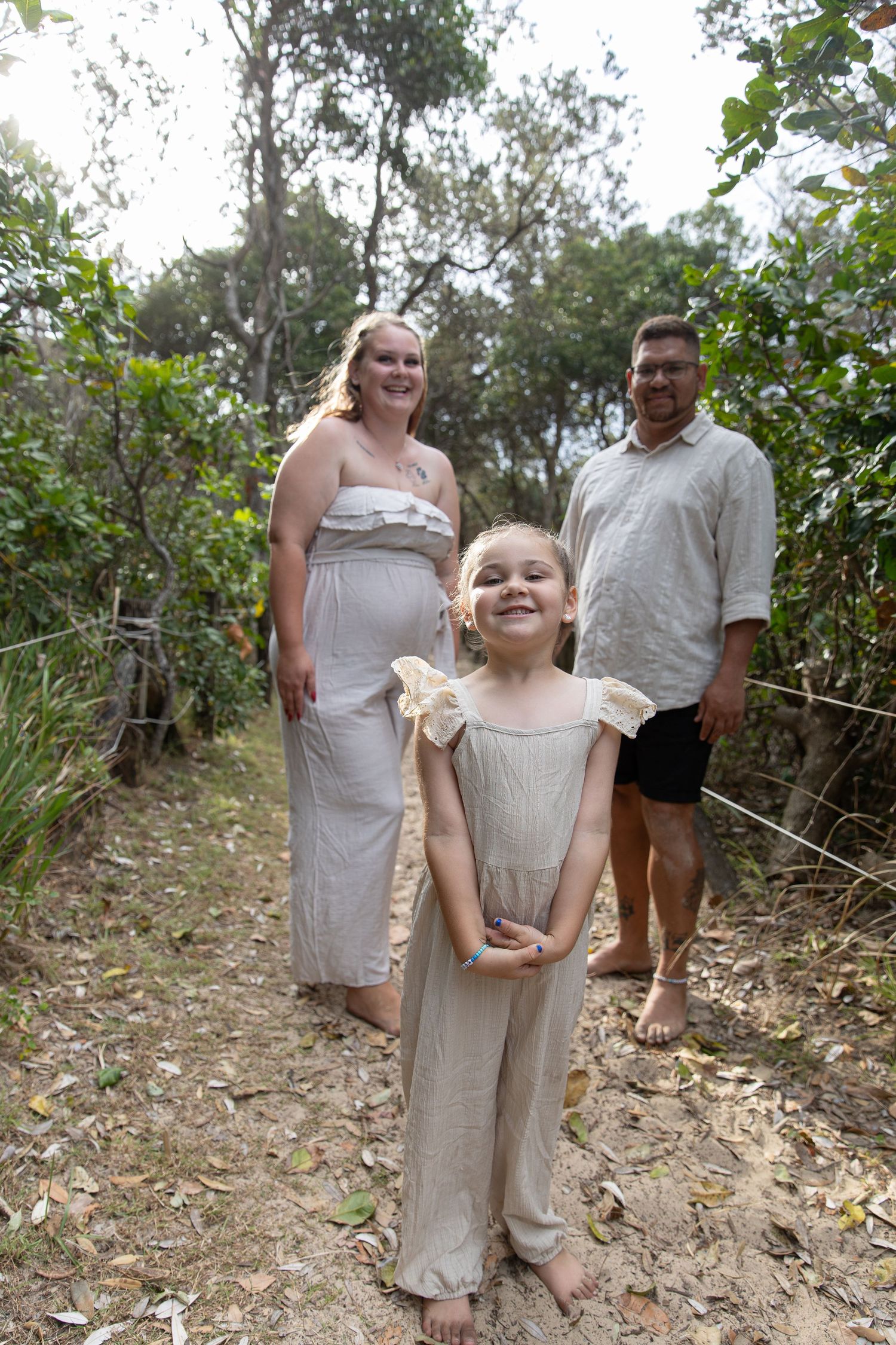 A pregnant woman and man stand behind a smiling young girl on a forest path, all wearing neutral linen outfits.
