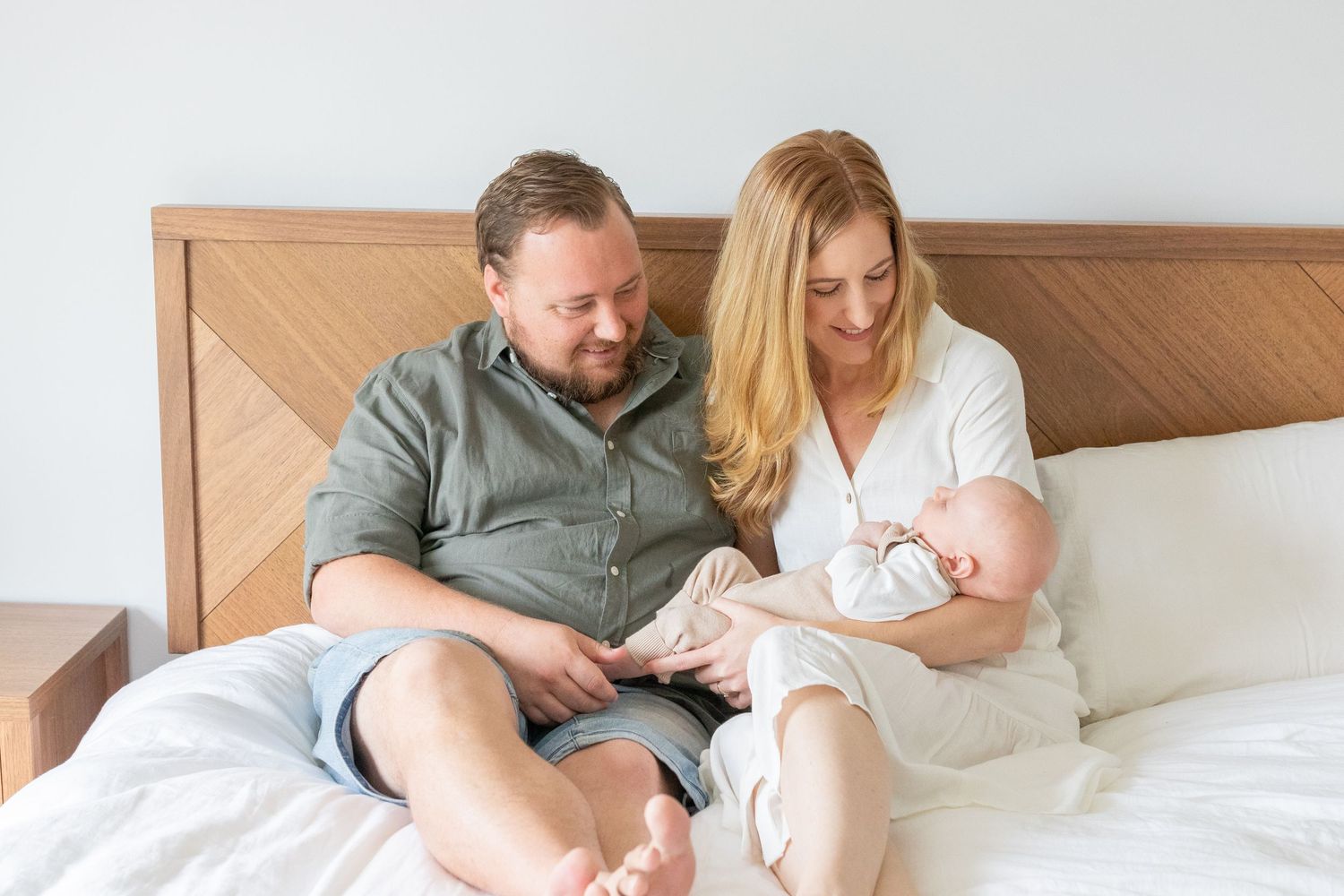 Happy couple sitting on bed admiring their newborn baby in a bright, cozy bedroom setting.