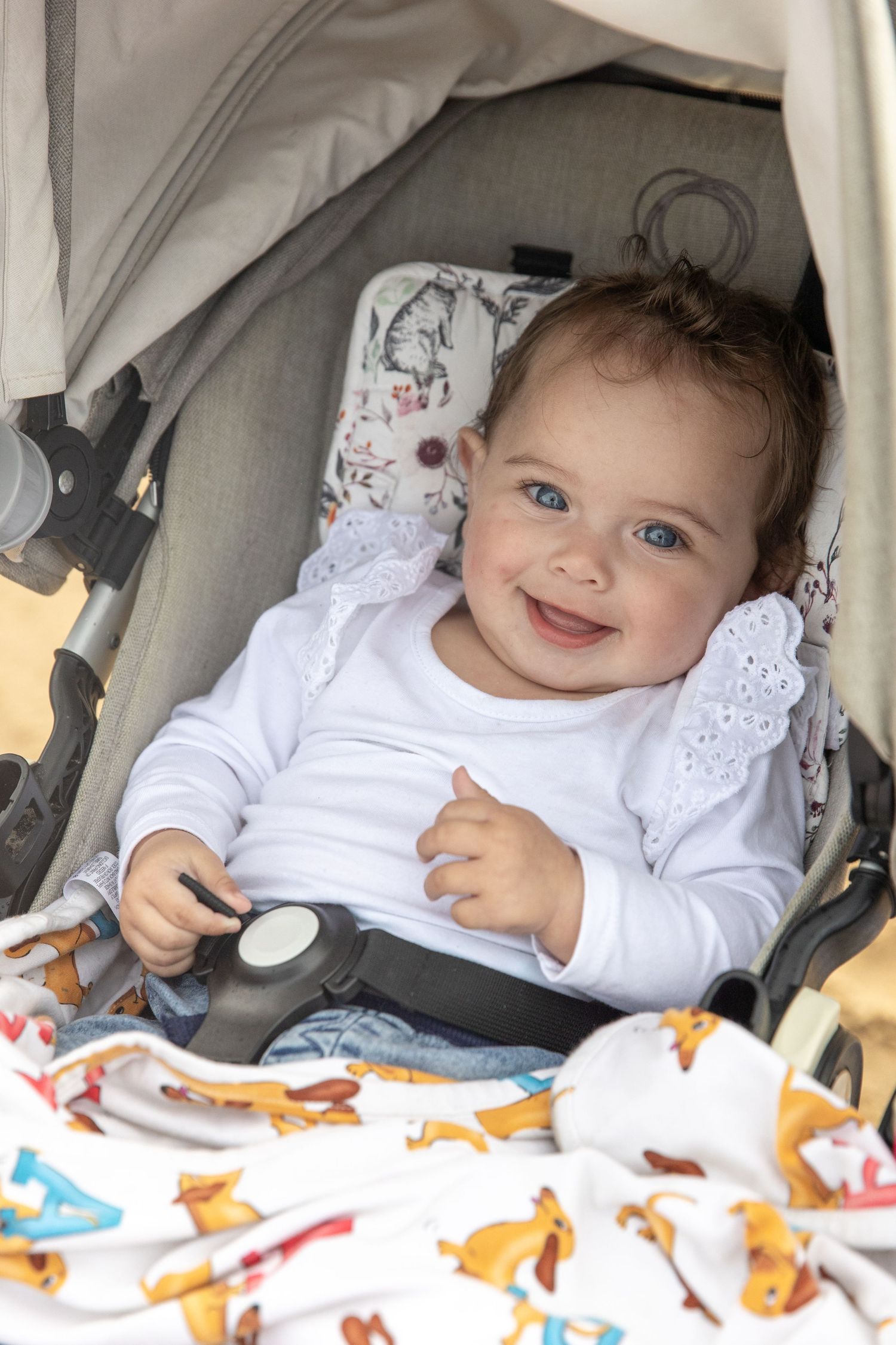 Happy baby with blue eyes smiling in a stroller, wearing a white outfit with a lace collar and colourful blanket. Coffs Coast photographer.