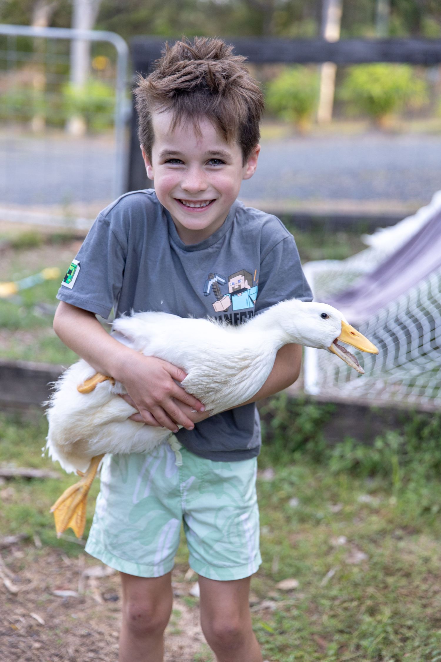 Child in grey shirt and mint shorts holding a white duck while smiling outdoors.