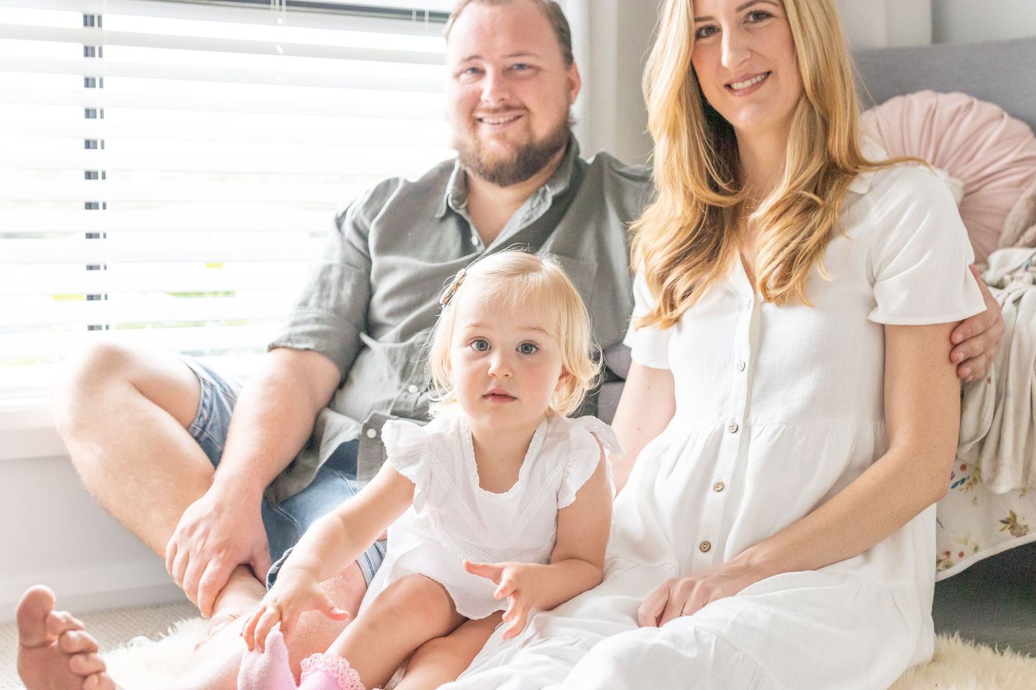 Happy family portrait with parents and toddler daughter sitting together by a bright window indoors.