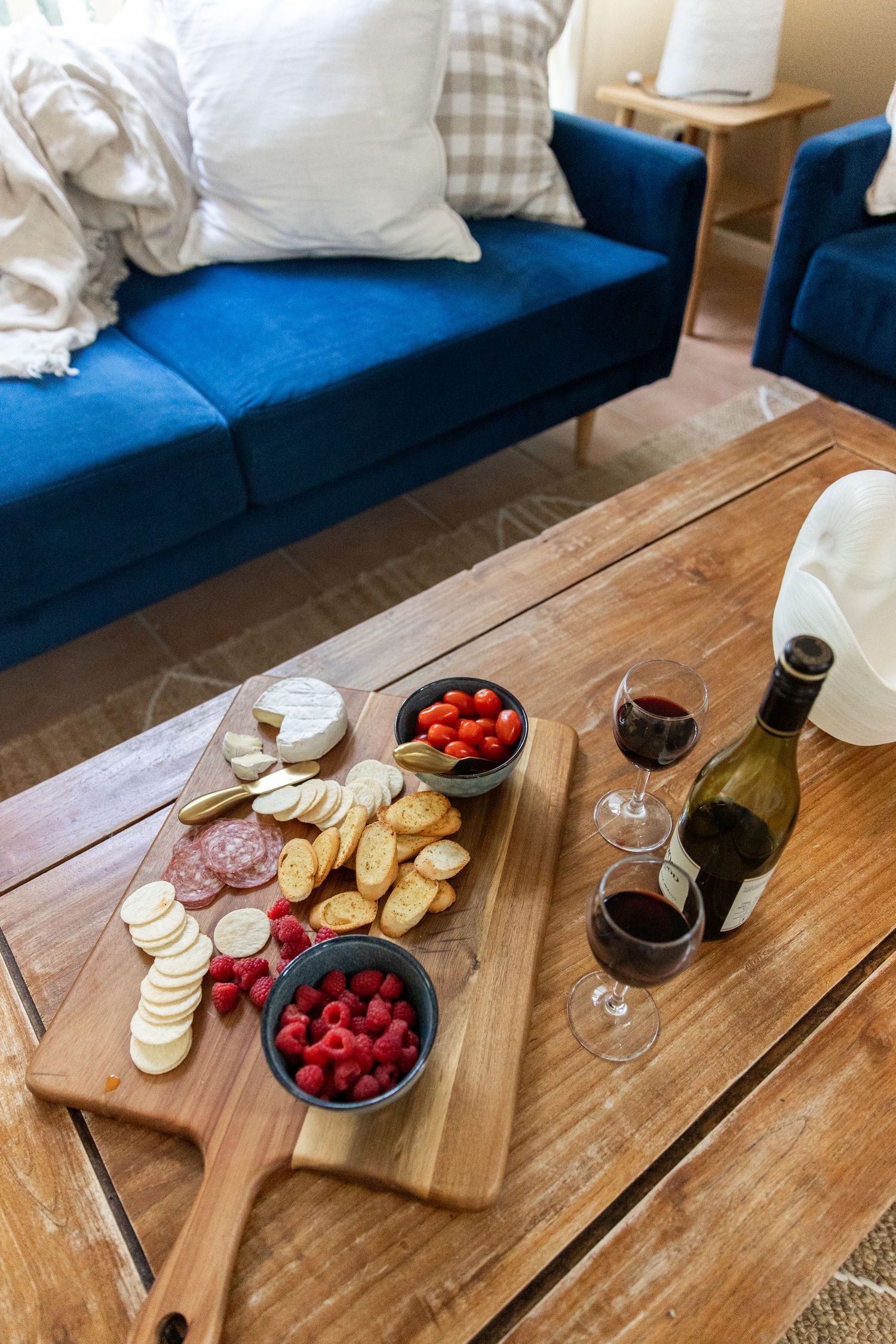 Charcuterie board with meats, cheeses, raspberries and wine glasses on a wooden coffee table.