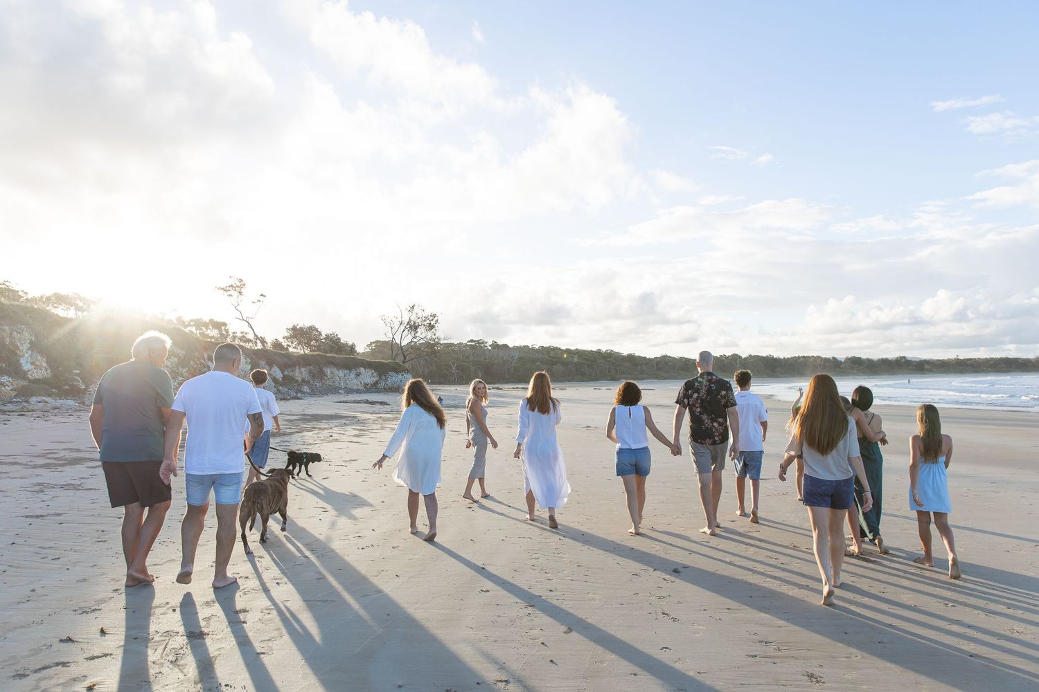 Large family and dog walking hand-in-hand along a sunny beach at sunset in white and blue outfits.