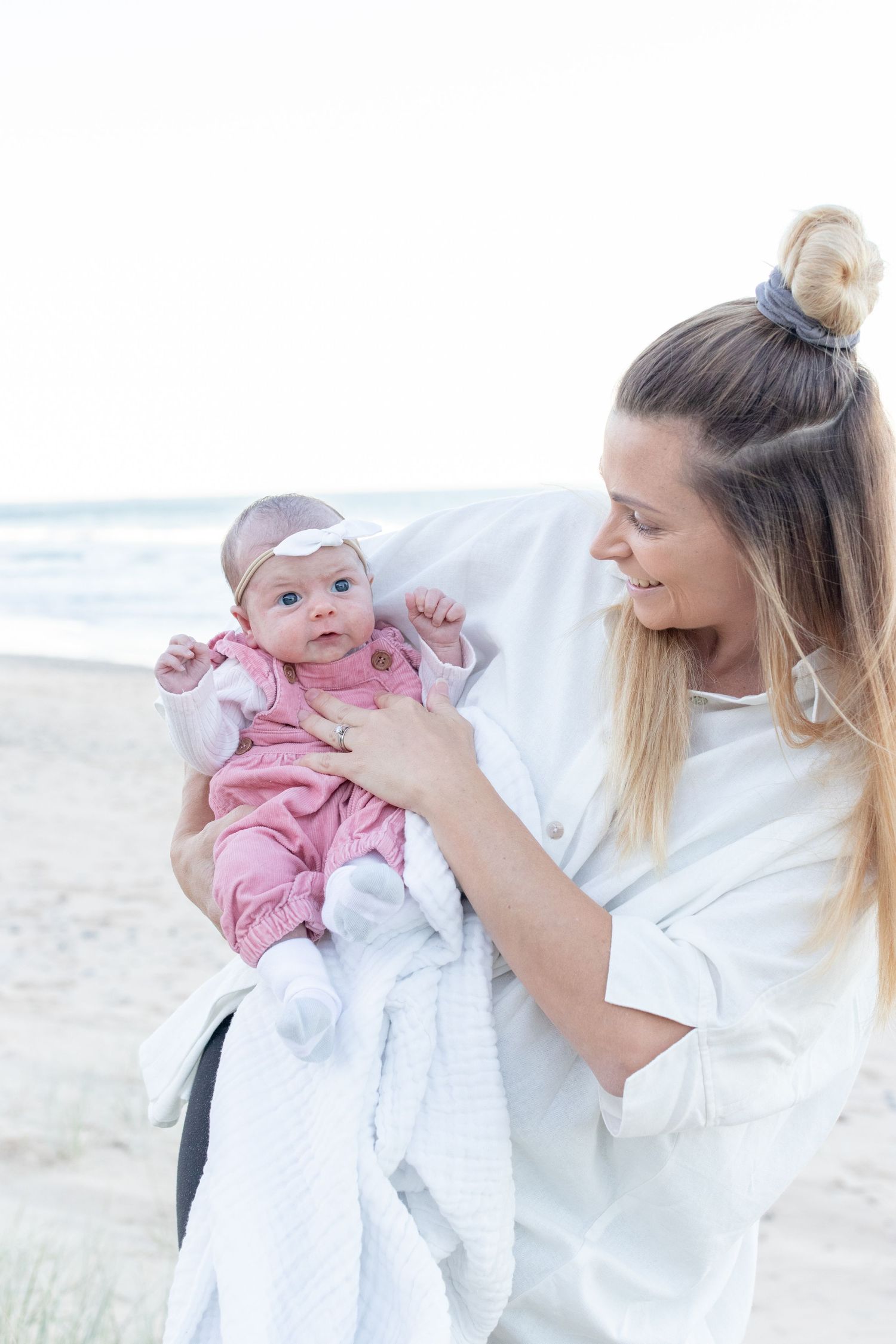 A smiling mother holds her newborn baby dressed in pink on a sandy beach. Coffs Coast photographer.